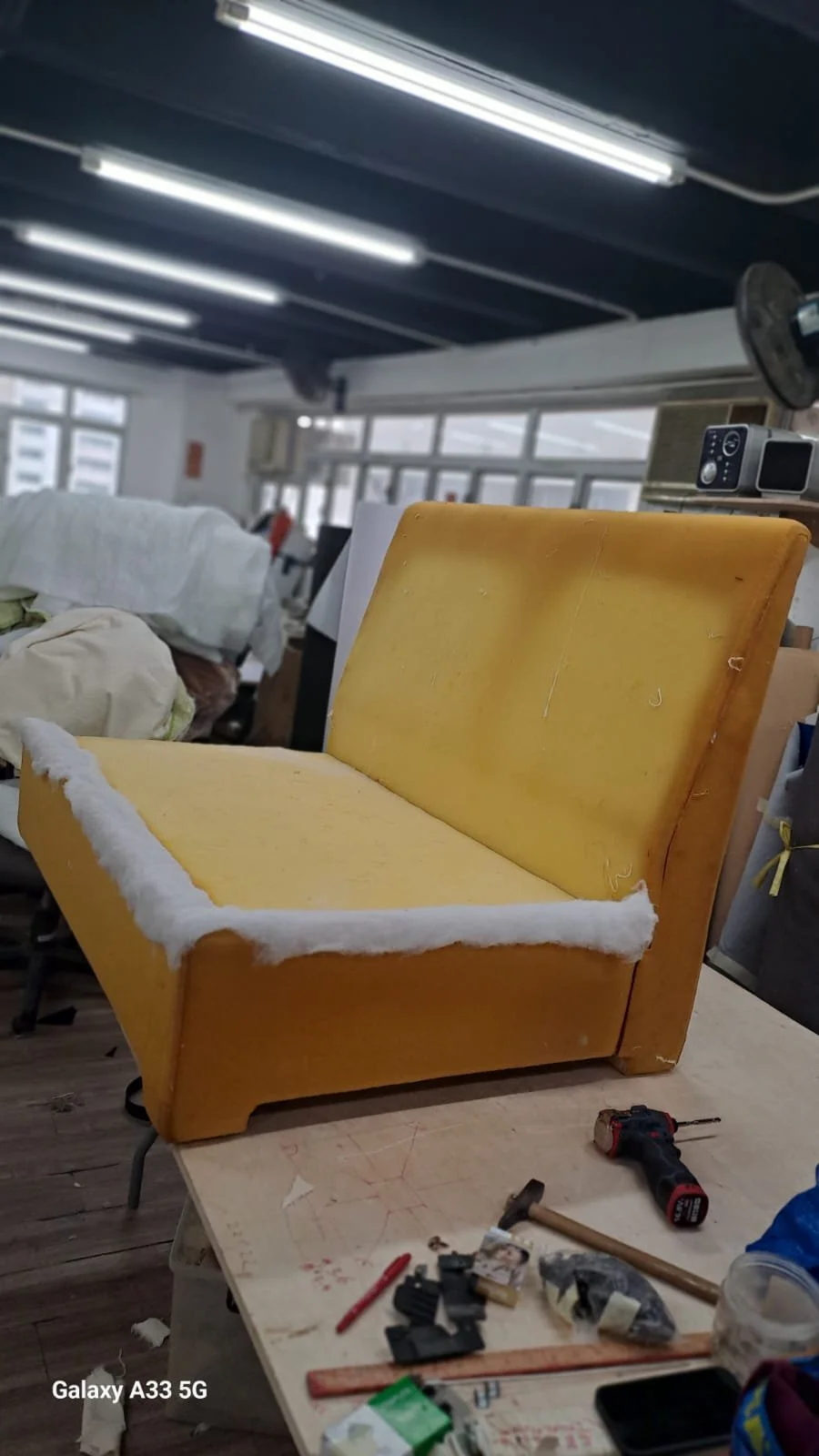 A yellow upholstered chair with one torn armrest and a piece of white fabric or stuffing on its seat, placed on a cluttered worktable in a workshop.
