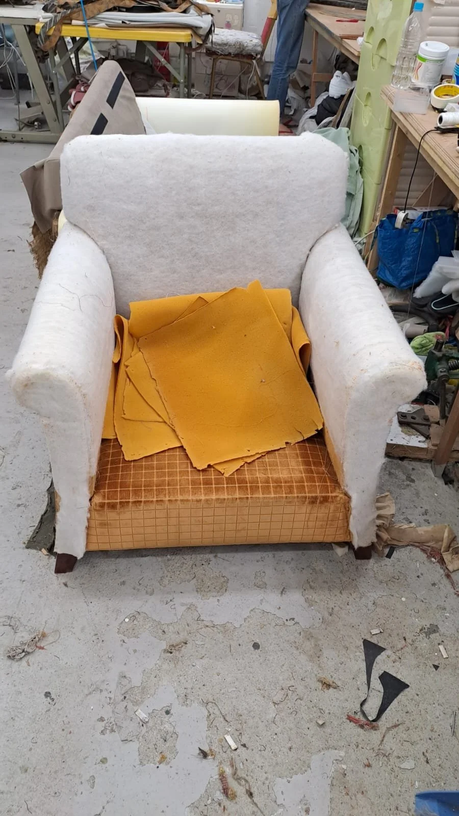 A worn, white upholstered armchair with a damaged seat cushion and a pile of yellow fabric or foam on top, in a cluttered workshop.