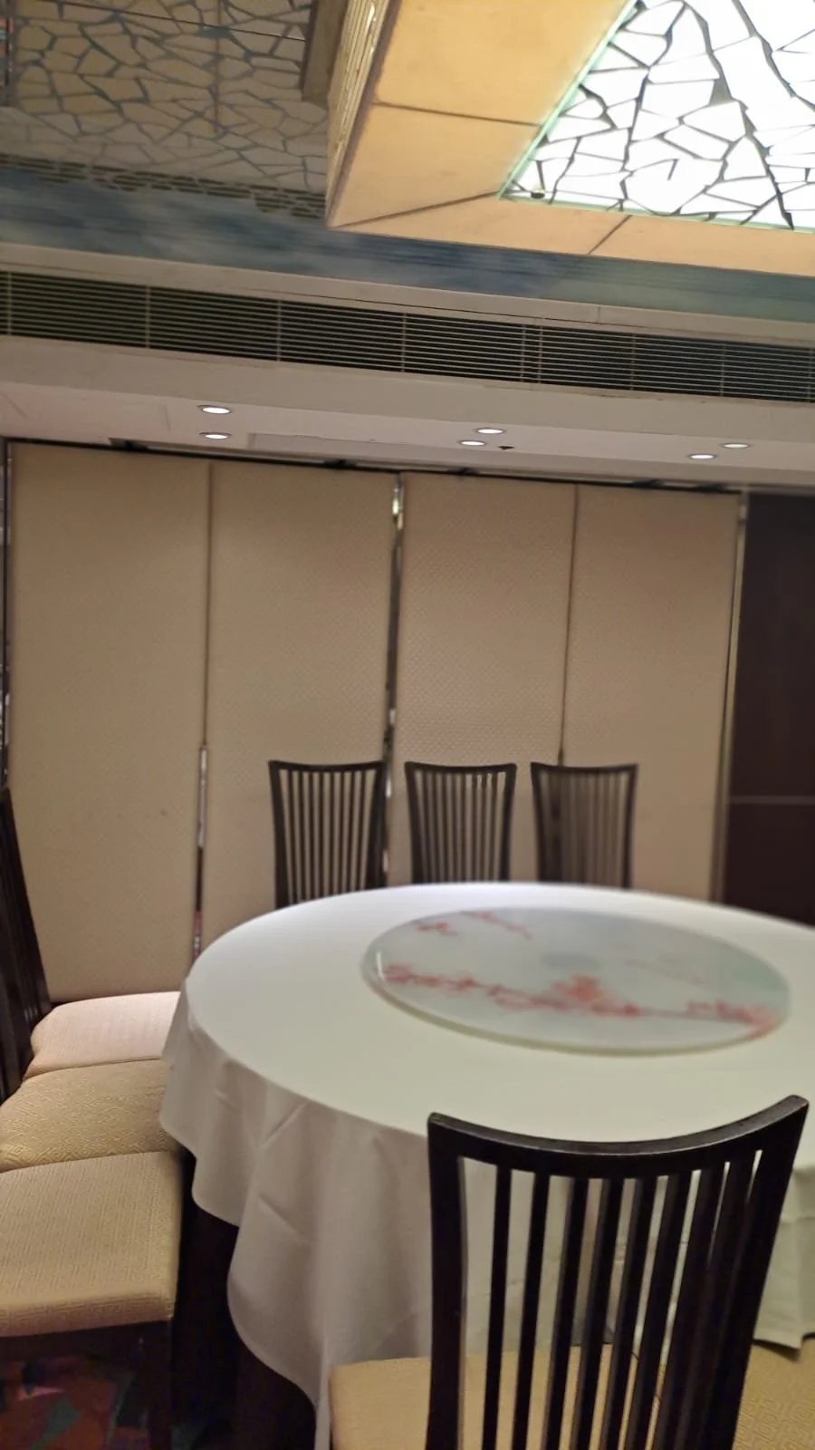 A round table with a white tablecloth and a glass Lazy Susan, surrounded by black wooden chairs in a dining room with beige folding partitions and a decorative ceiling.