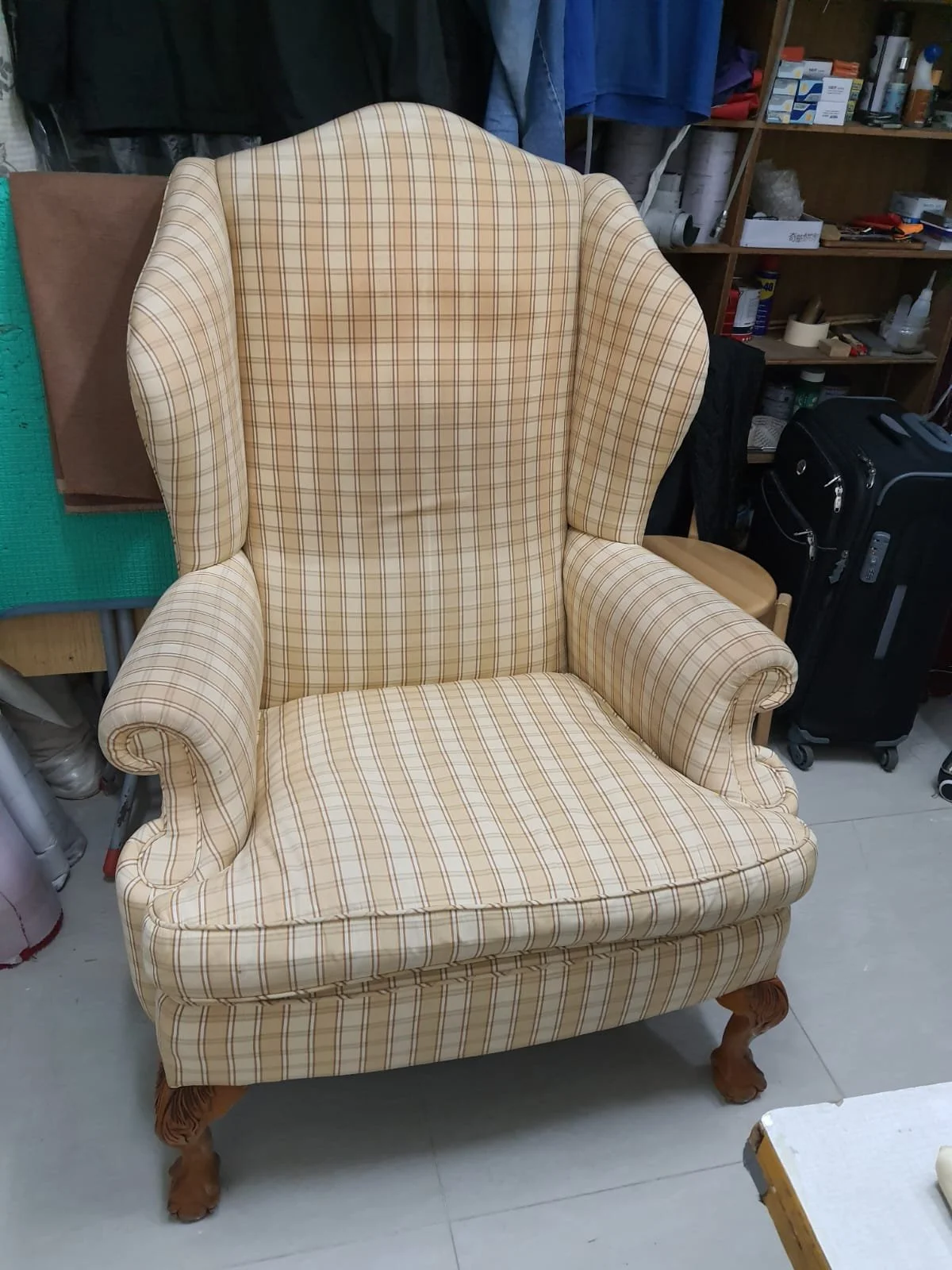 A beige armchair with a plaid pattern and wooden clawfoot legs, placed in a cluttered room with shelves and boxes in the background.