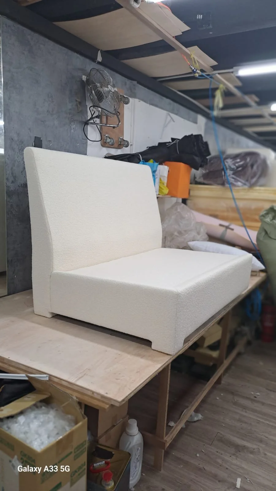 A cream-colored upholstered chair on a wooden workbench in a workshop surrounded by various tools, boxes, and materials.