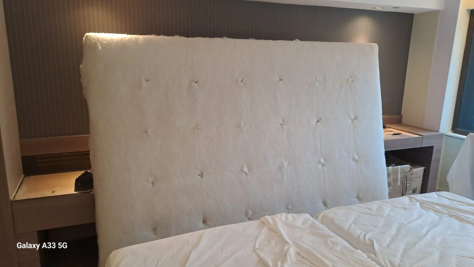 A hotel room featuring a large, cream-colored tufted headboard, partially obscuring the bed. There are side tables on either side, with some items on the left side table. To the right, there's a desk with a window beside it, letting in natural light.