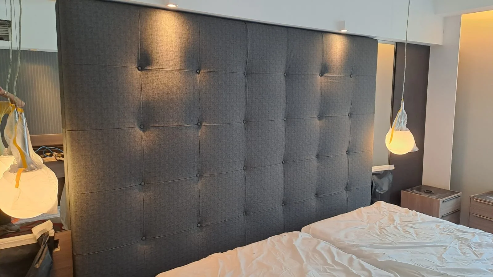 A hotel room with a large, padded, dark gray headboard behind two unmade beds with white sheets. There are two hanging lamps with white shades on either side of the headboard. A nightstand is visible on the right, holding a tissue box and other small items. The left side of the room shows a cluttered surface with a bag or plastic wrapping hanging from the lamp.