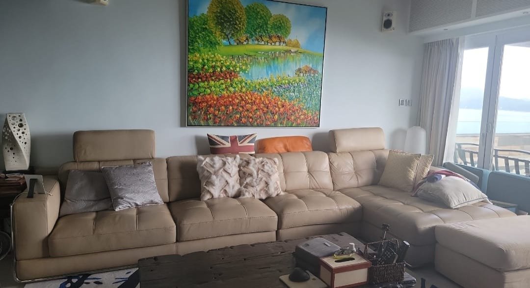 A living room with a beige sectional sofa decorated with multiple pillows, a colorful landscape painting hanging on the wall, a wooden coffee table with various items, and a large window with a sliding door leading to a balcony.