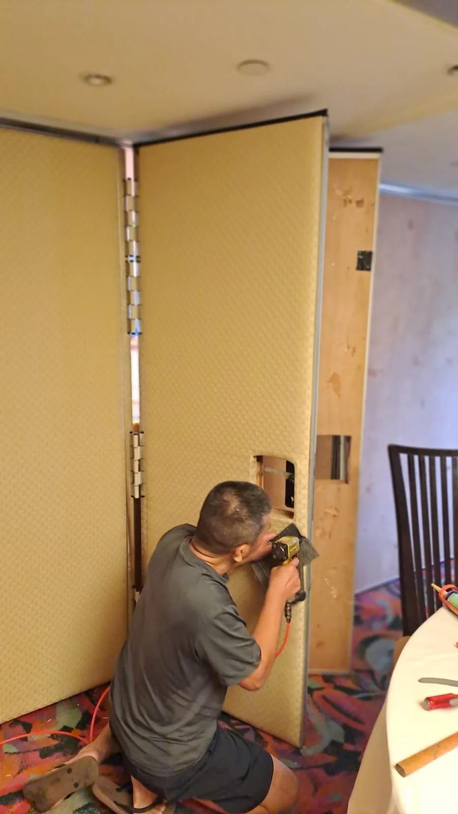 A man working on a construction or renovation project, using a power drill on a panel or wall in a room with a patterned carpet and a dining table nearby.