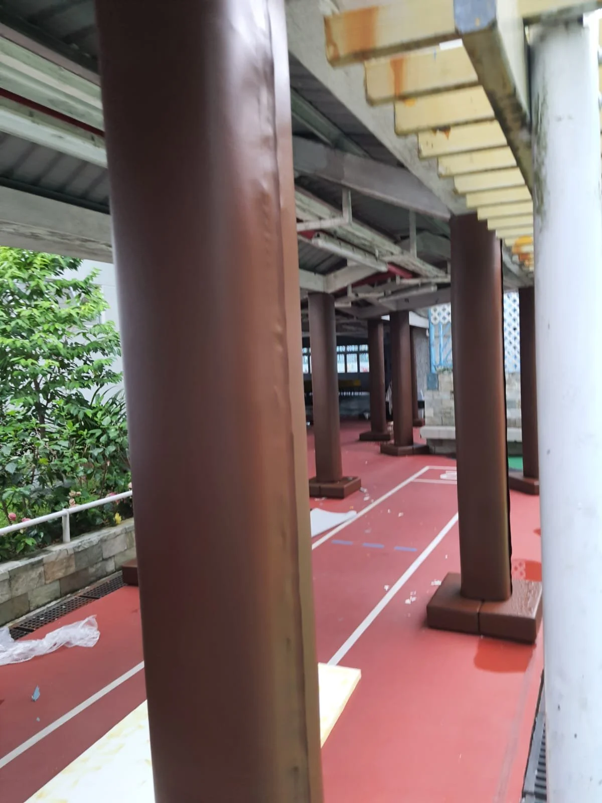 View of a covered outdoor seating area with brown wooden pillars, a red concrete floor, some painted lines, and a garden with green plants on the side.