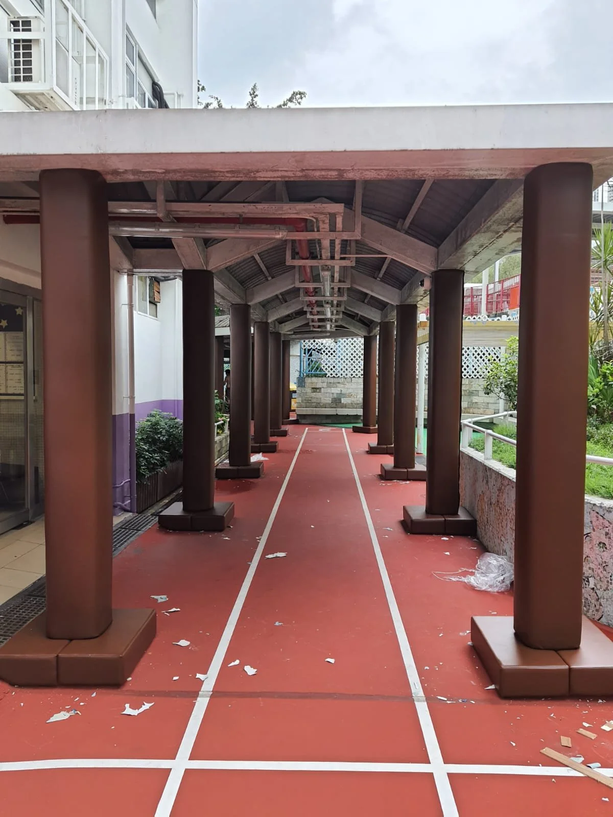 Covered outdoor walkway with brown columns and a red floor, with white lines resembling a running track.