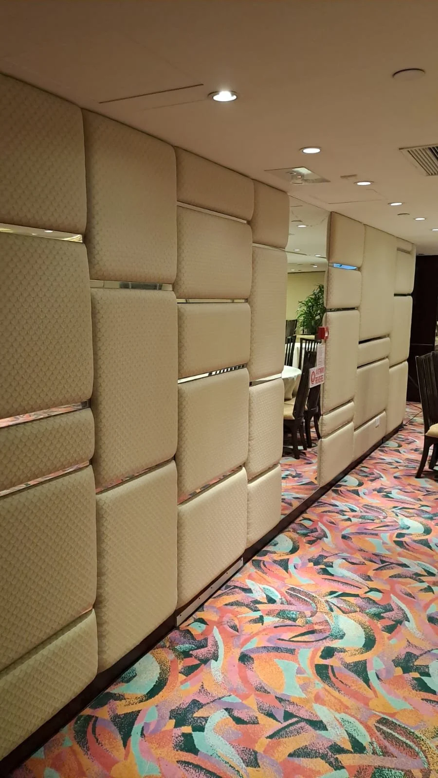 Interior of a hotel or restaurant with padded beige partition walls, patterned carpet, and dining tables and chairs in the background.