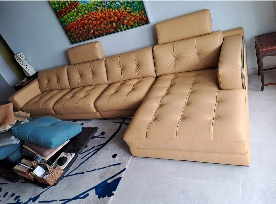 A tan L-shaped leather sofa in a living room with a colorful painting on the wall behind it. There is a black coffee table with books, a blue pillow, and a brown blanket on it. A small side table with a decorative item is beside the sofa.
