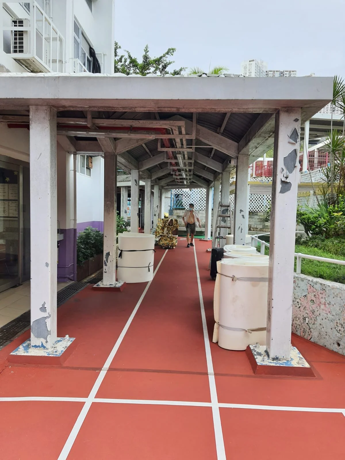 View of a walkway with an open-sided roof, red flooring with white lines, stacked white chairs or tables, a person walking away, and greenery to the side.