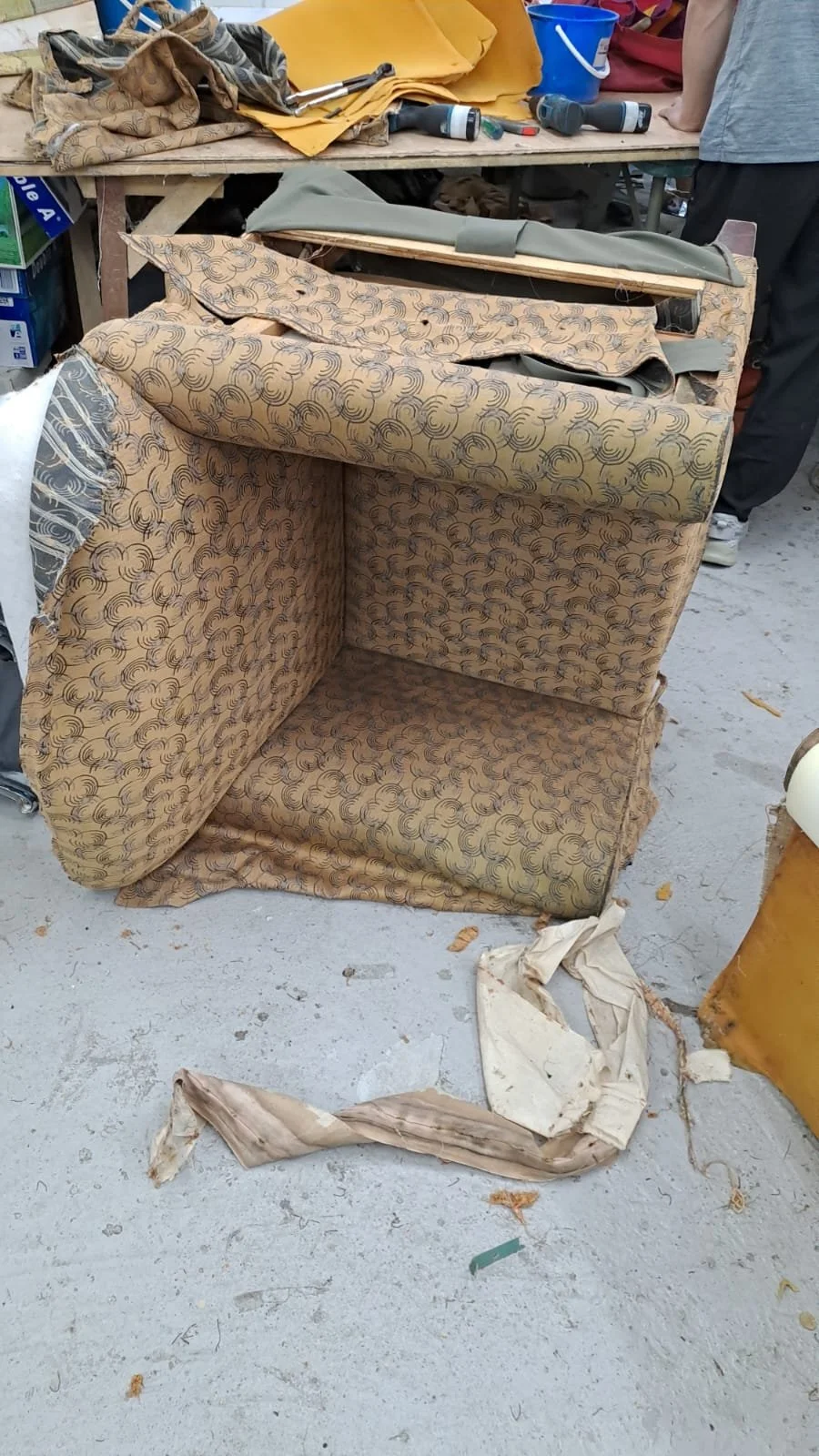 A partially disassembled upholstered furniture piece with a patterned fabric, sitting on a rough concrete floor in a cluttered workspace. Items like fabric scraps, tools, and other work supplies are visible on a table behind it.