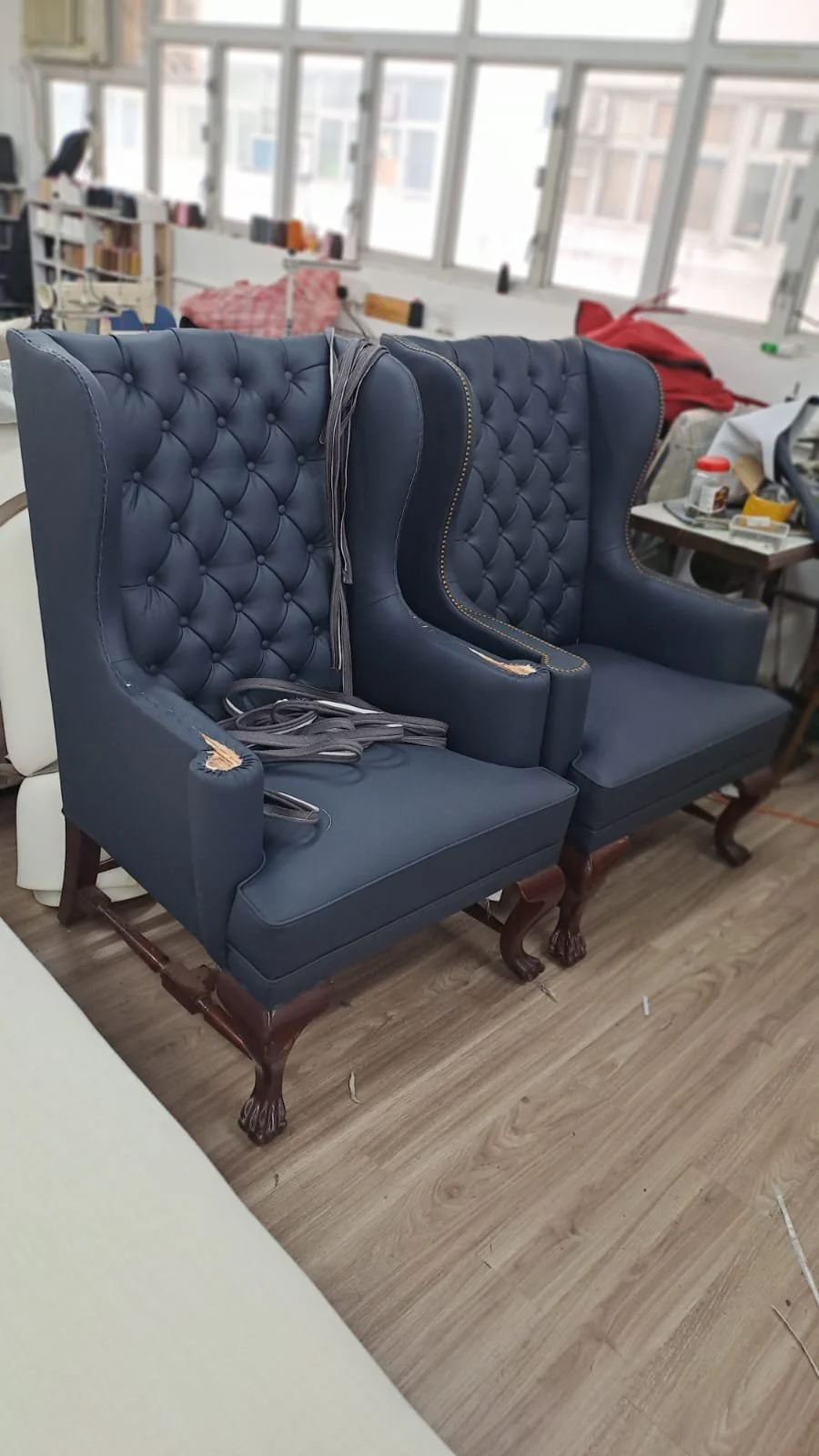 Two navy blue high-back tufted armchairs with wooden carved legs in a cluttered room, with miscellaneous items and a large window in the background.