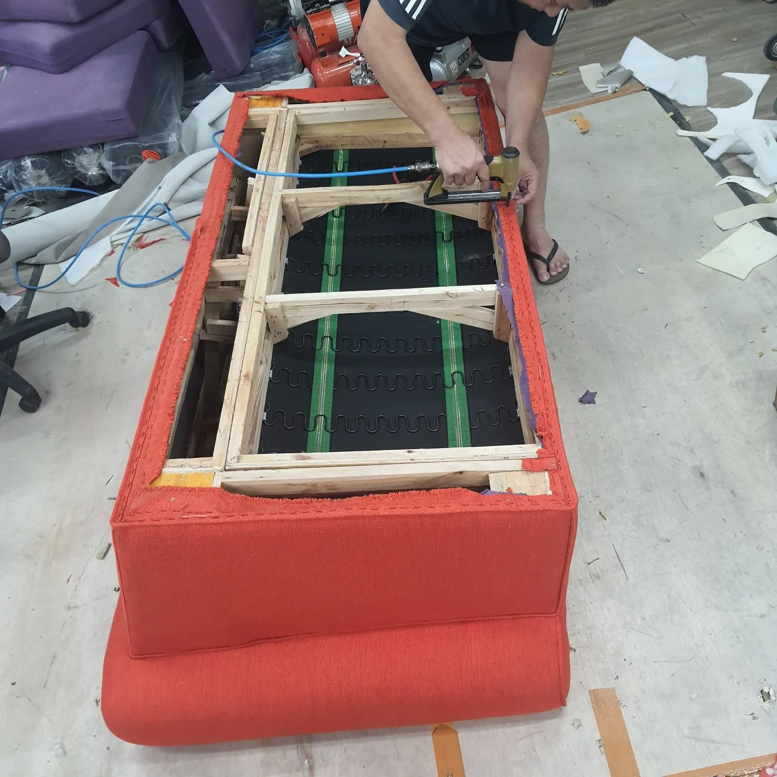 Person upholstering a red sofa with a wooden frame removed, working with a staple gun in a workshop surrounded by fabric scraps and tools.