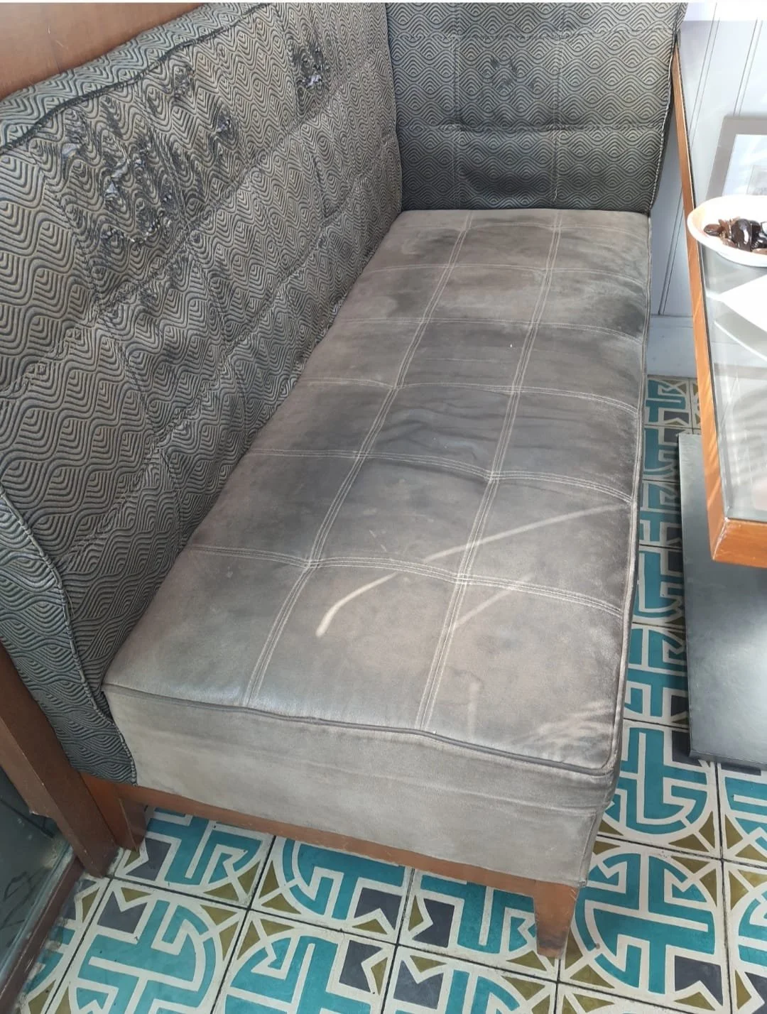 A vintage sofa with a patterned backrest and a plain seat cushion. The sofa is placed on a colorful patterned tile floor, next to a glass-topped table with a white plate holding various items.