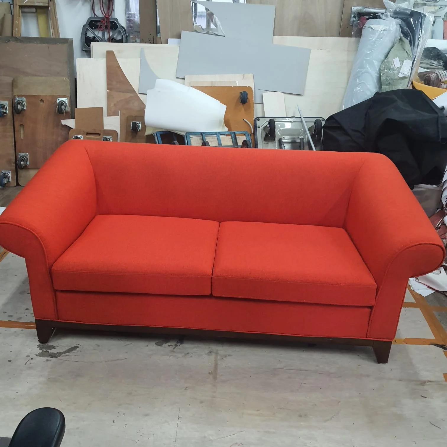 A red upholstered sofa with rolled arms and wooden legs, placed in a workshop or storage area with various office supplies and materials in the background.