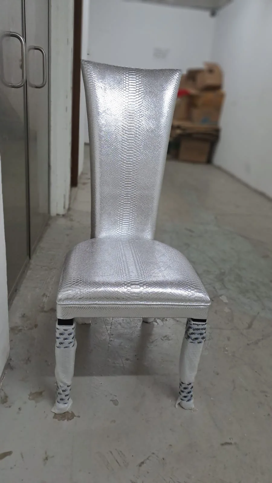 A silver, metallic, textured dining chair with wrapped legs in bubble wrap, located in a room with a concrete floor and cardboard boxes in the background.