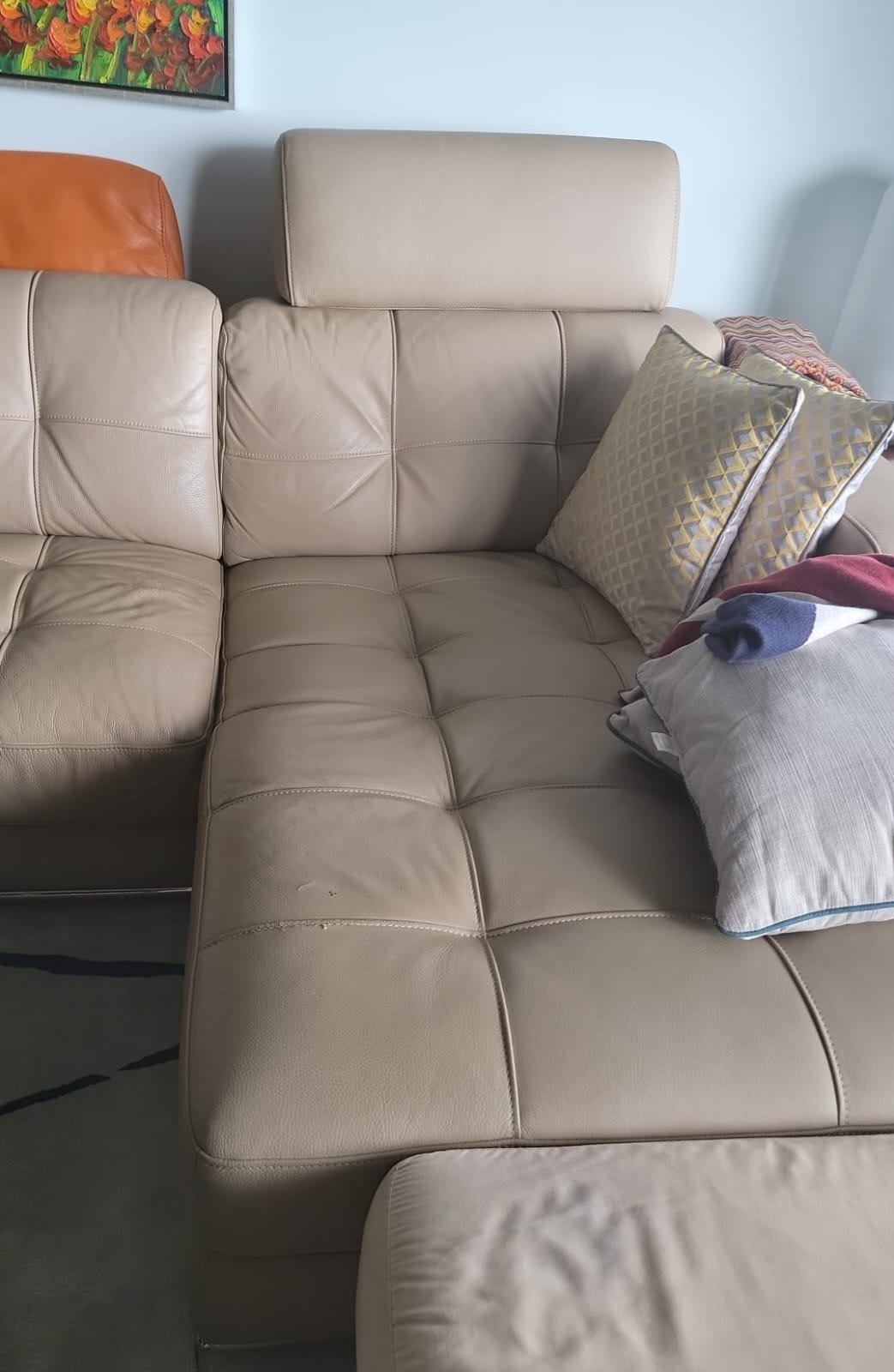 A beige leather sectional sofa with tufted cushions and a matching headrest. The sofa has several pillows, including a decorative pillow with a geometric pattern. There is a colorful painting on the wall above the sofa. Part of an orange leather chai