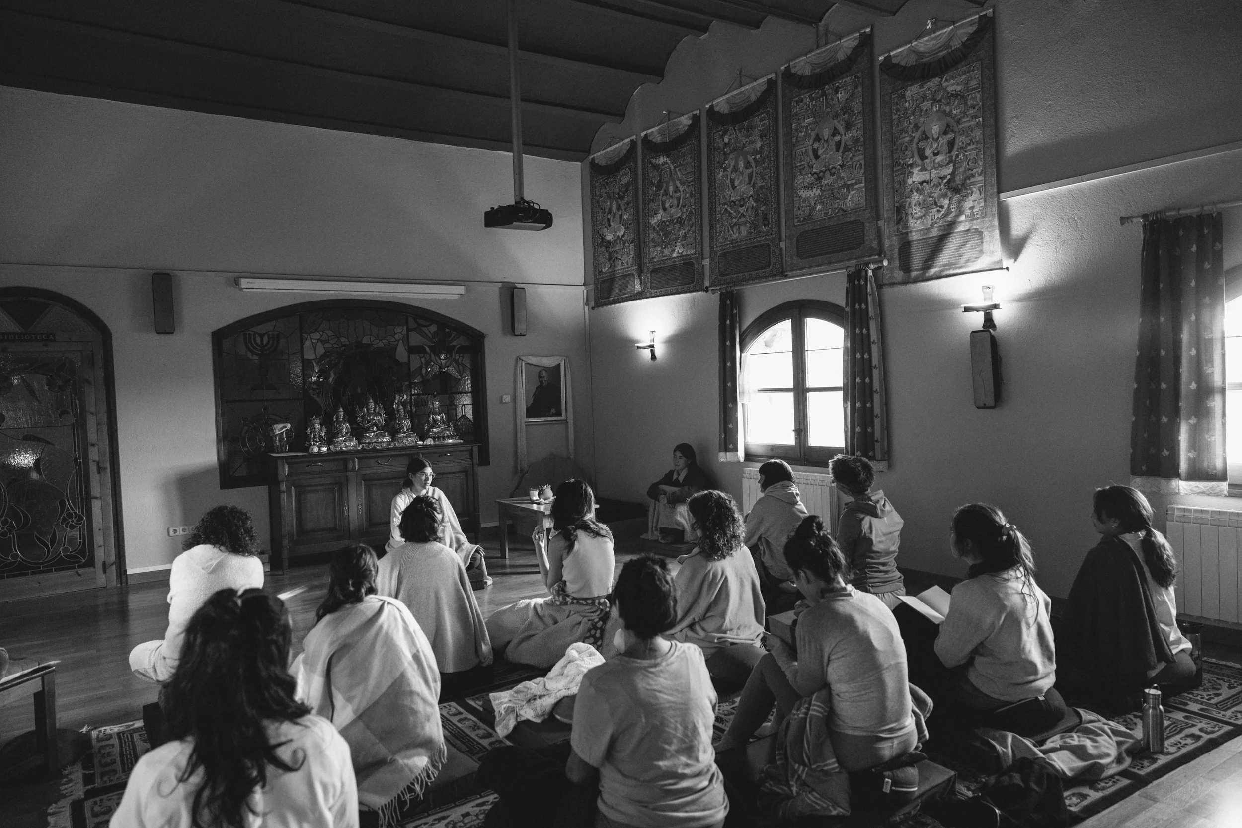 Grupo de personas sentadas en el suelo en una sala, participando en una actividad o ceremonia, con una figura de Buda y arte religioso en el fondo, luz que entra por las ventanas.