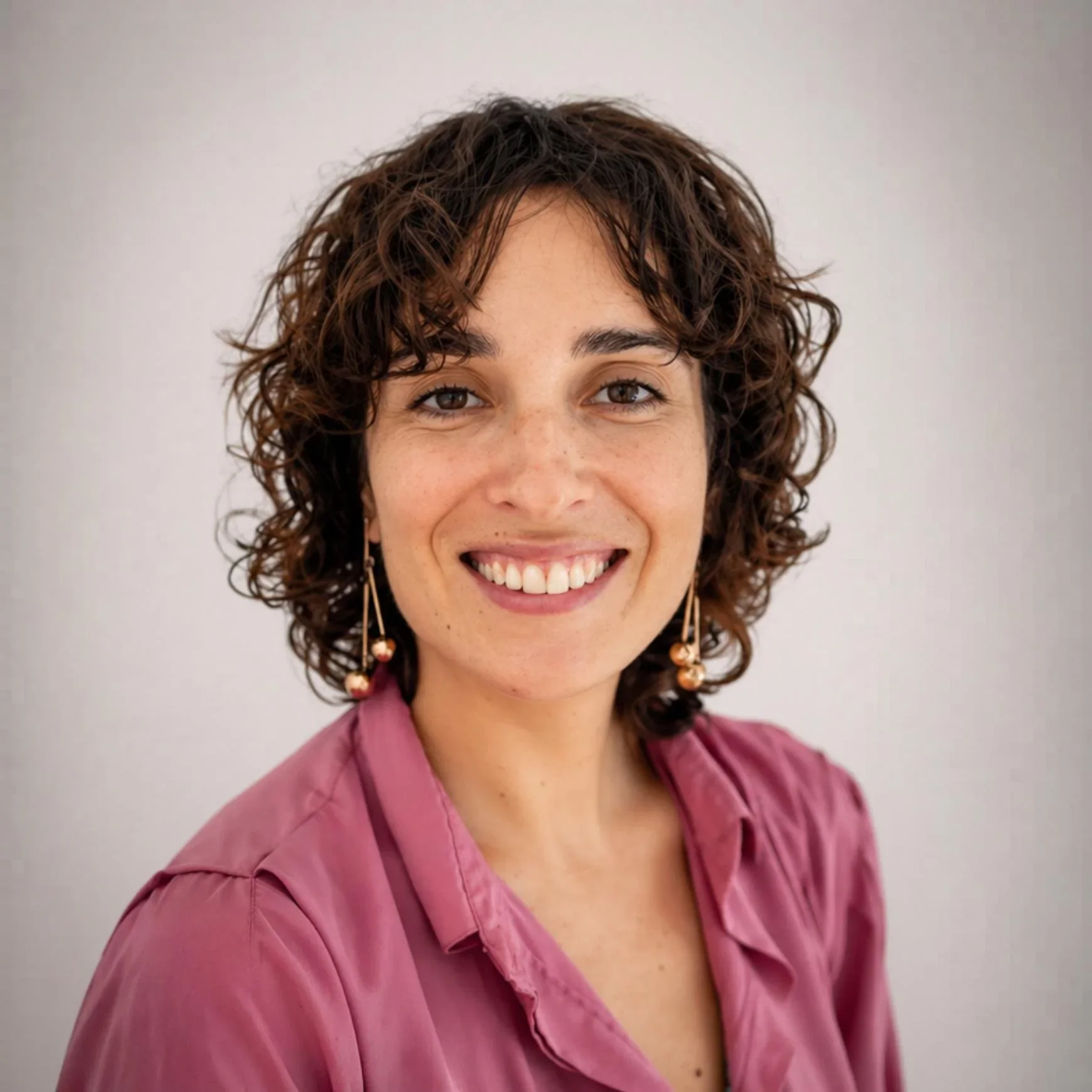 Retrato de una mujer sonriente con cabello rizado y corto, usando blusa rosa y pendientes de arete largo, frente a fondo gris.