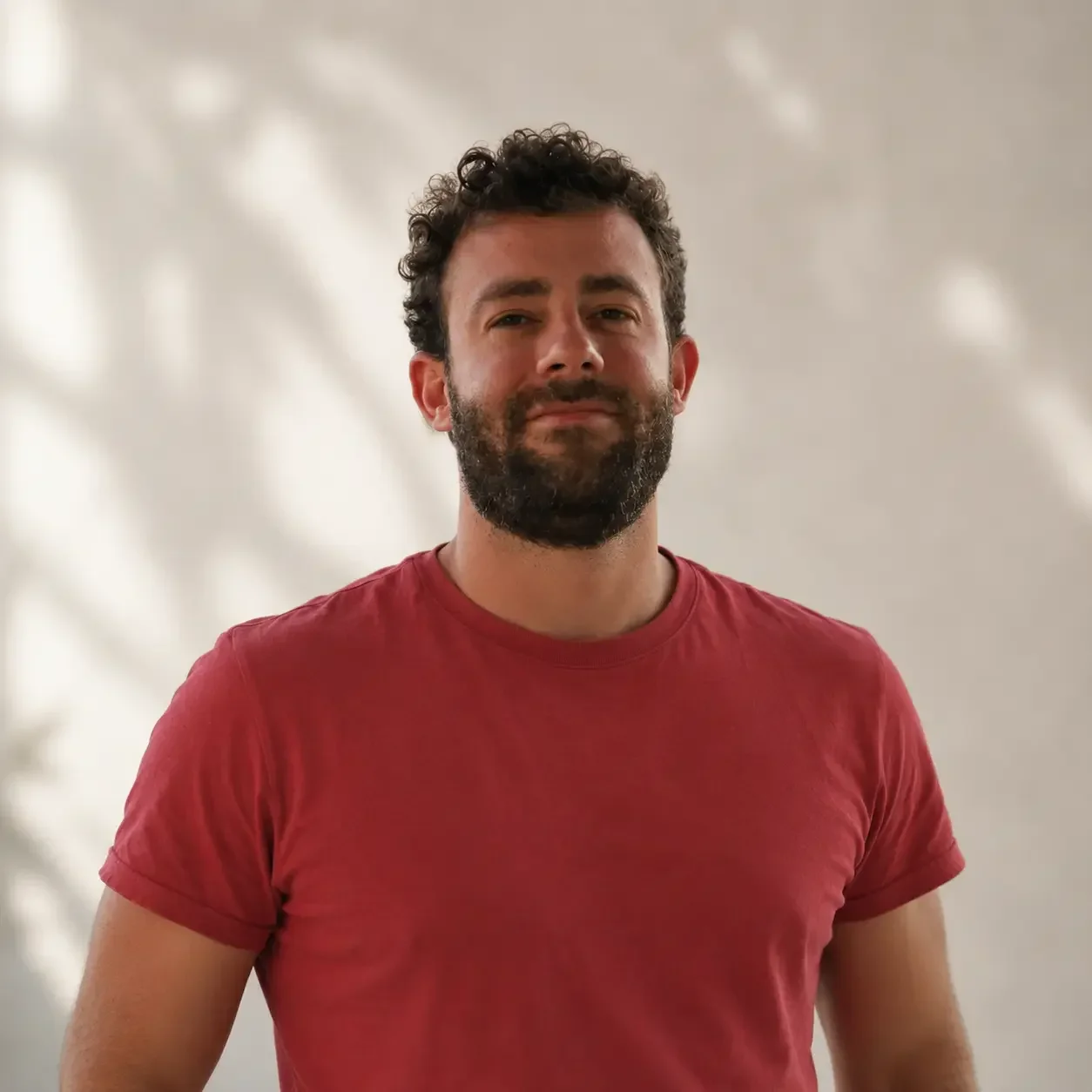 Hombre con barba, cabello rizado y camiseta roja en un fondo difuso.