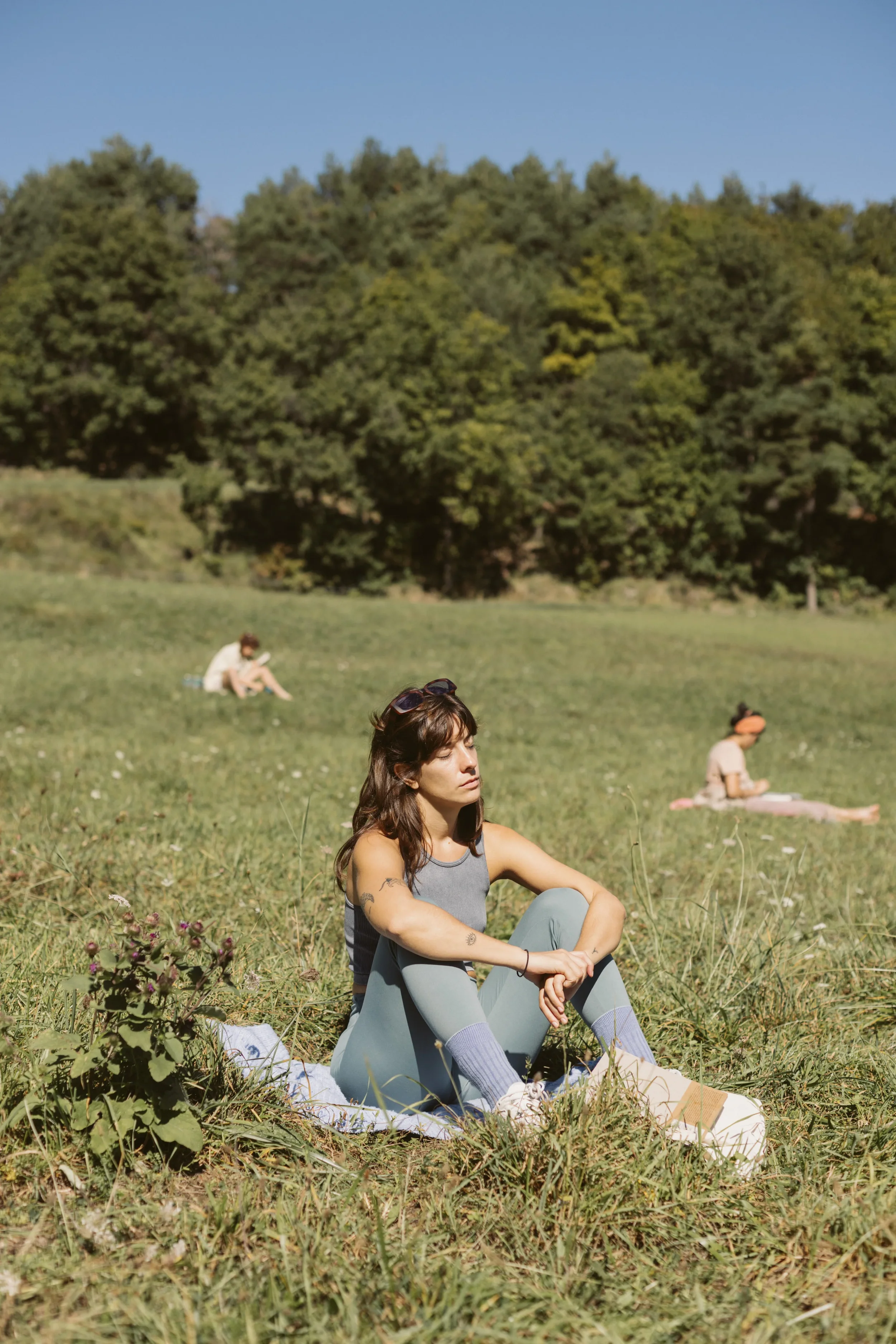 Mujer con cabello castaño medianamente rizado, brazos tatuados, sentada en la hierba, con un conjunto de ropa deportiva gris, rodeada de otras personas en fondo, en un paisaje verde con árboles y cielo azul.