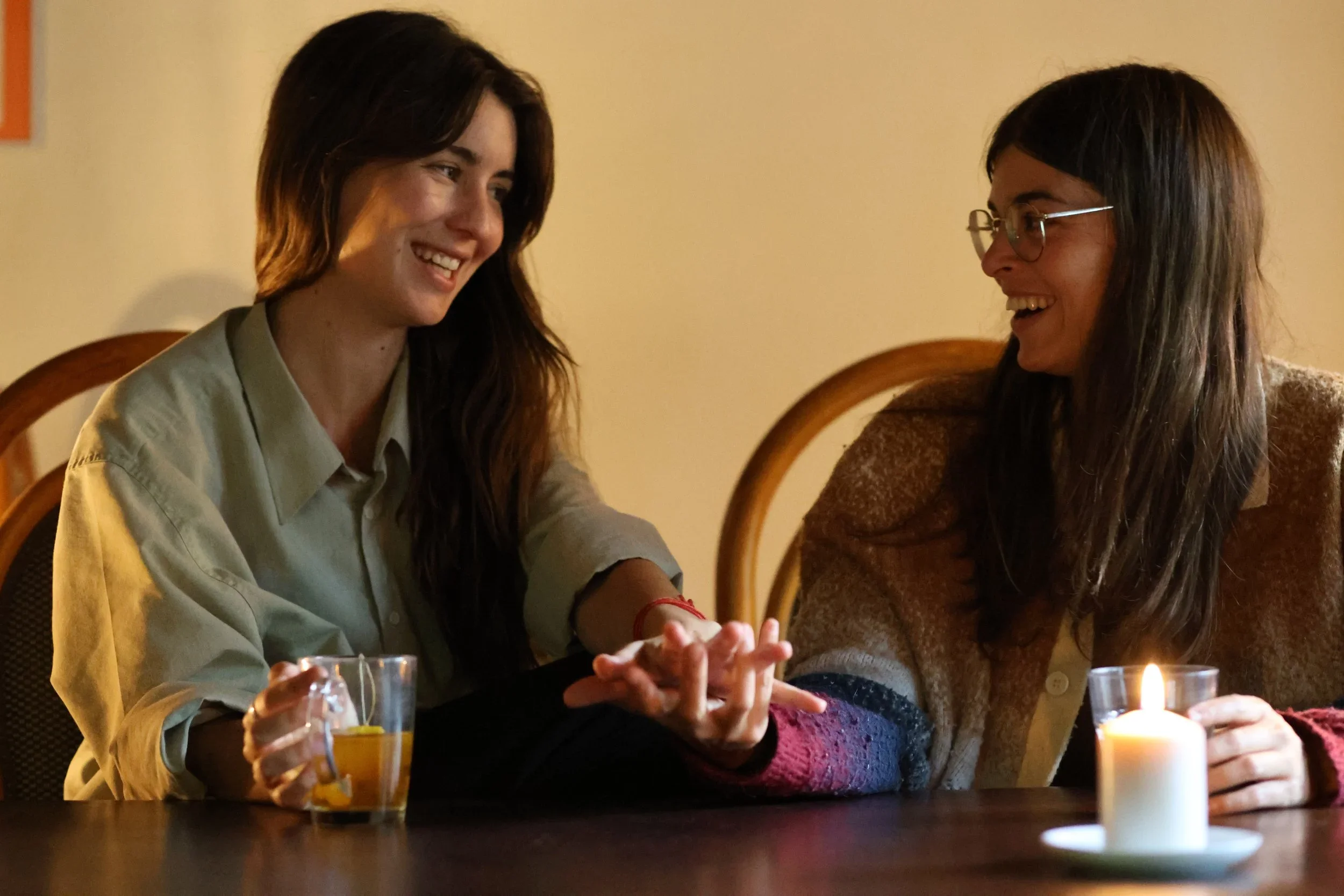 Dos mujeres conversando y sonriendo en una mesa, con velas y bebidas en primer plano.