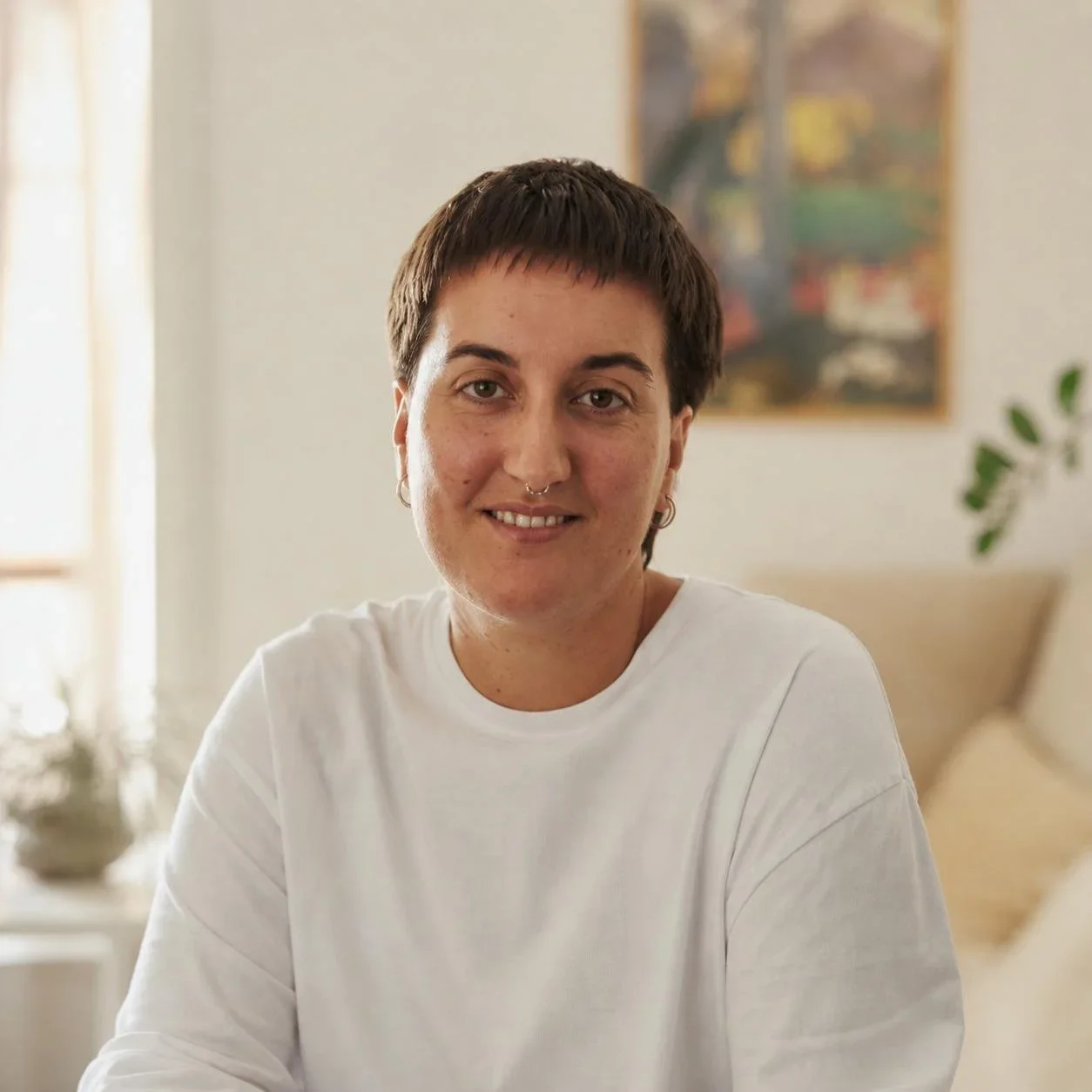 Persona con cabello corto y oscuro, usando camiseta blanca, sonriendo en un ambiente luminoso y acogedor con decoraciones en un fondo difuso.