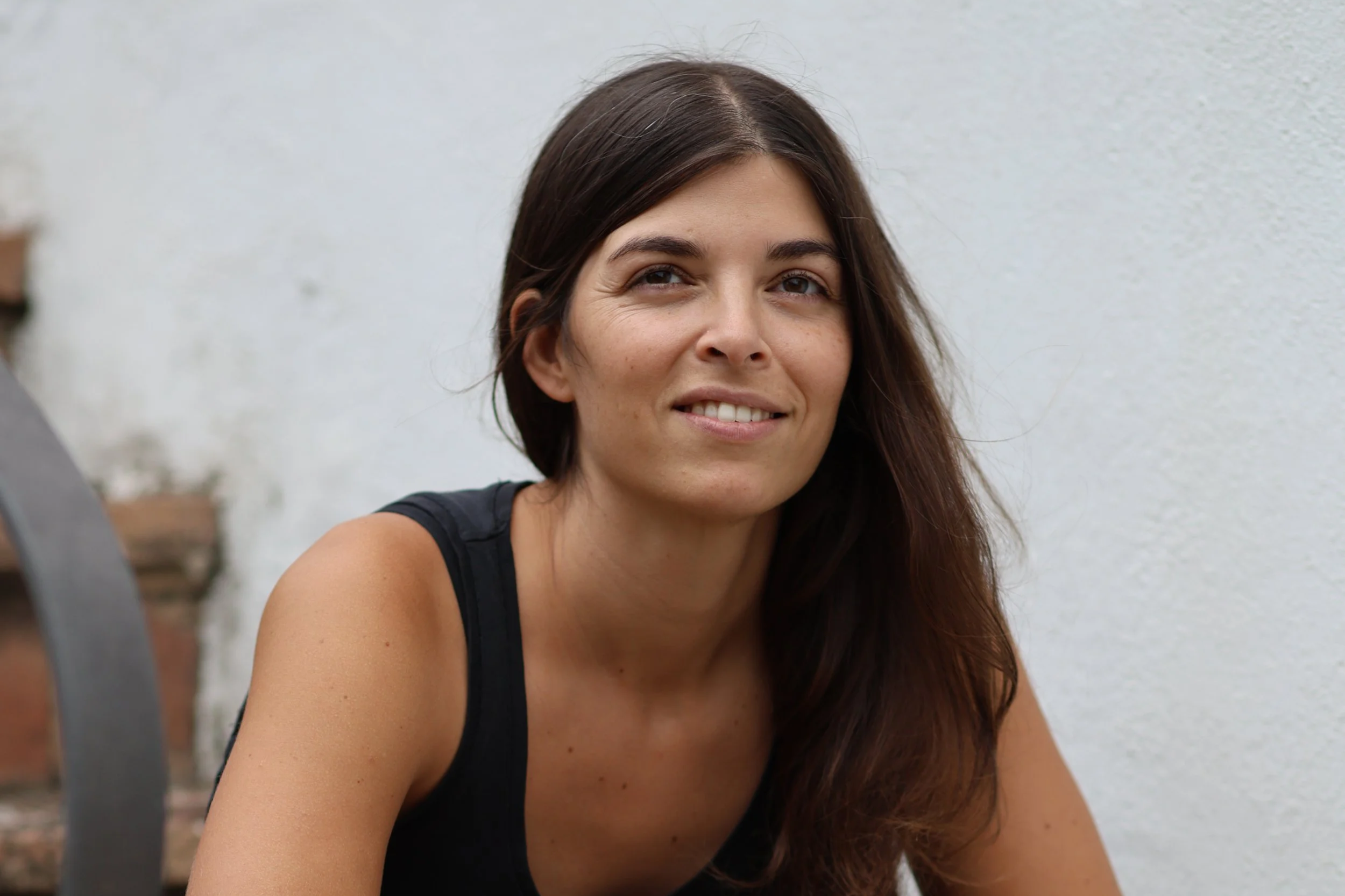 Mujer de piel morena con cabello largo y oscuro, sonriendo ligeramente, usando un top negro, en un fondo de muro blanco y elementos de ladrillo y madera en un ambiente exterior.