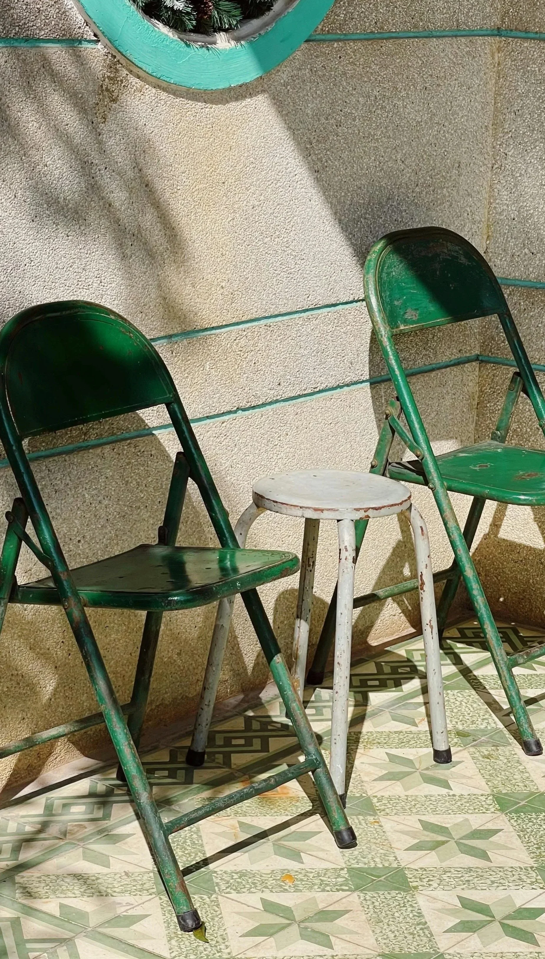 Tres sillas viejas y oxidadas, una de ellas blanca y las otras verdes, en un espacio cerrado con paredes de textura rugosa y piso con mosaico decorativo en tonos verdes y beige.