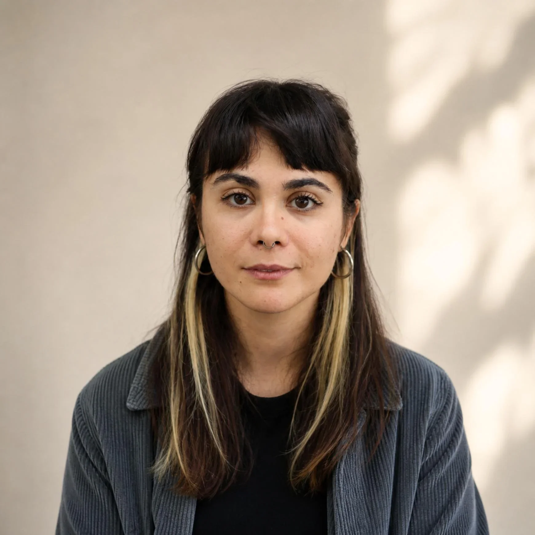 Retrato de una mujer joven con pelo oscuro y mechones rubios, usando aretes grandes y un suéter a rayas, con fondo beige y sombra en la pared.