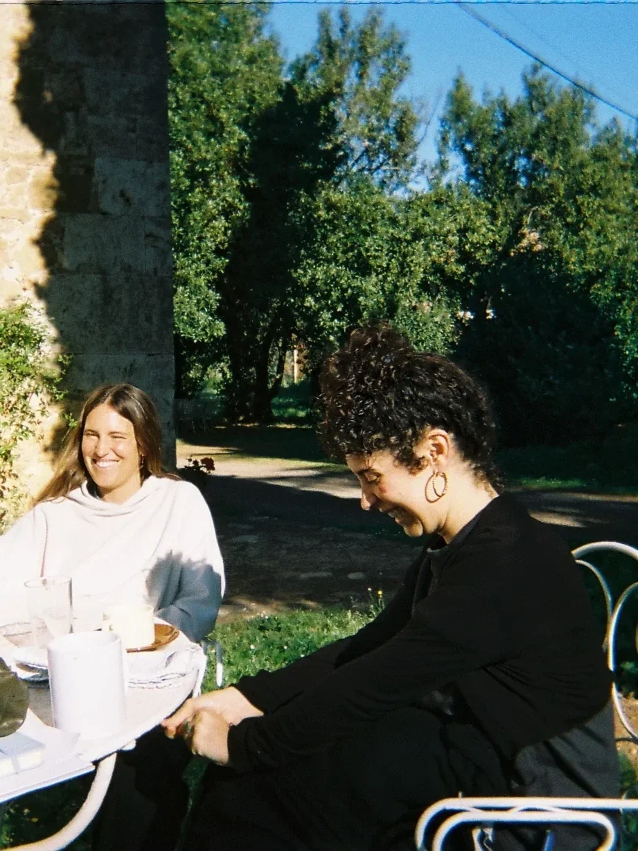 Dos mujeres sonriendo y riendo en una mesa al aire libre con platos y vasos, rodeadas de árboles y plantas verdes bajo un cielo azul.