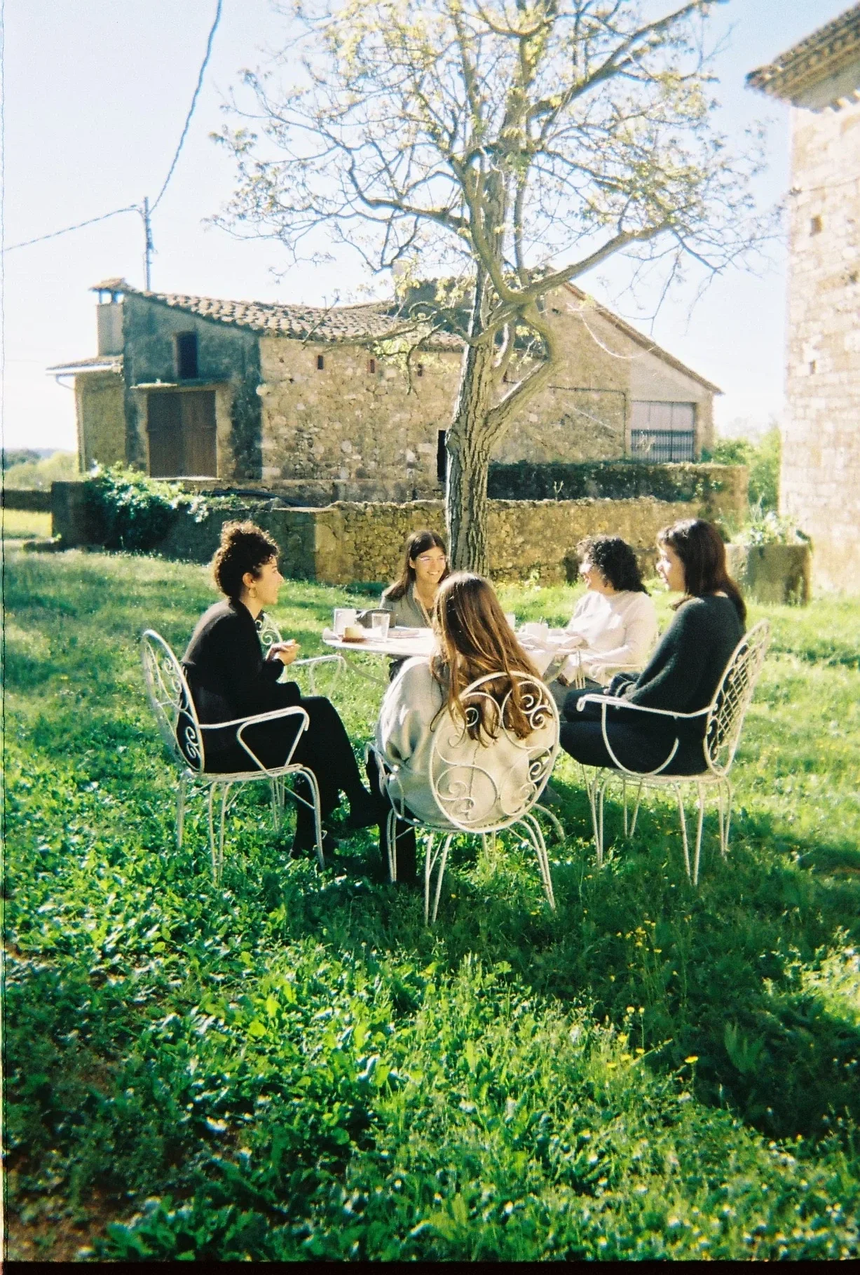 Grupo de cinco mujeres sentadas en sillas blancas de metal en un jardín con césped, disfrutando de una conversación, con árboles y una casa antigua de piedra en el fondo y un día soleado.