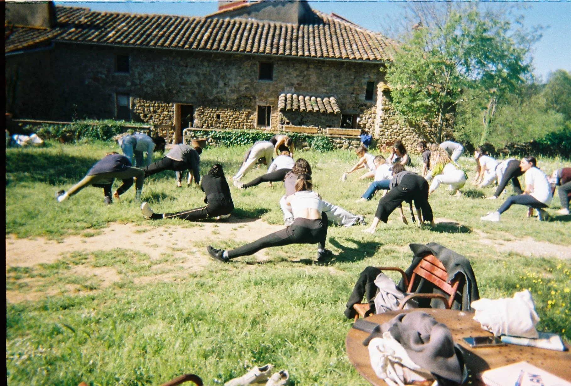 Grupo de personas realizando ejercicio de estiramiento en un parque frente a una casa de piedra con techo de tejas.