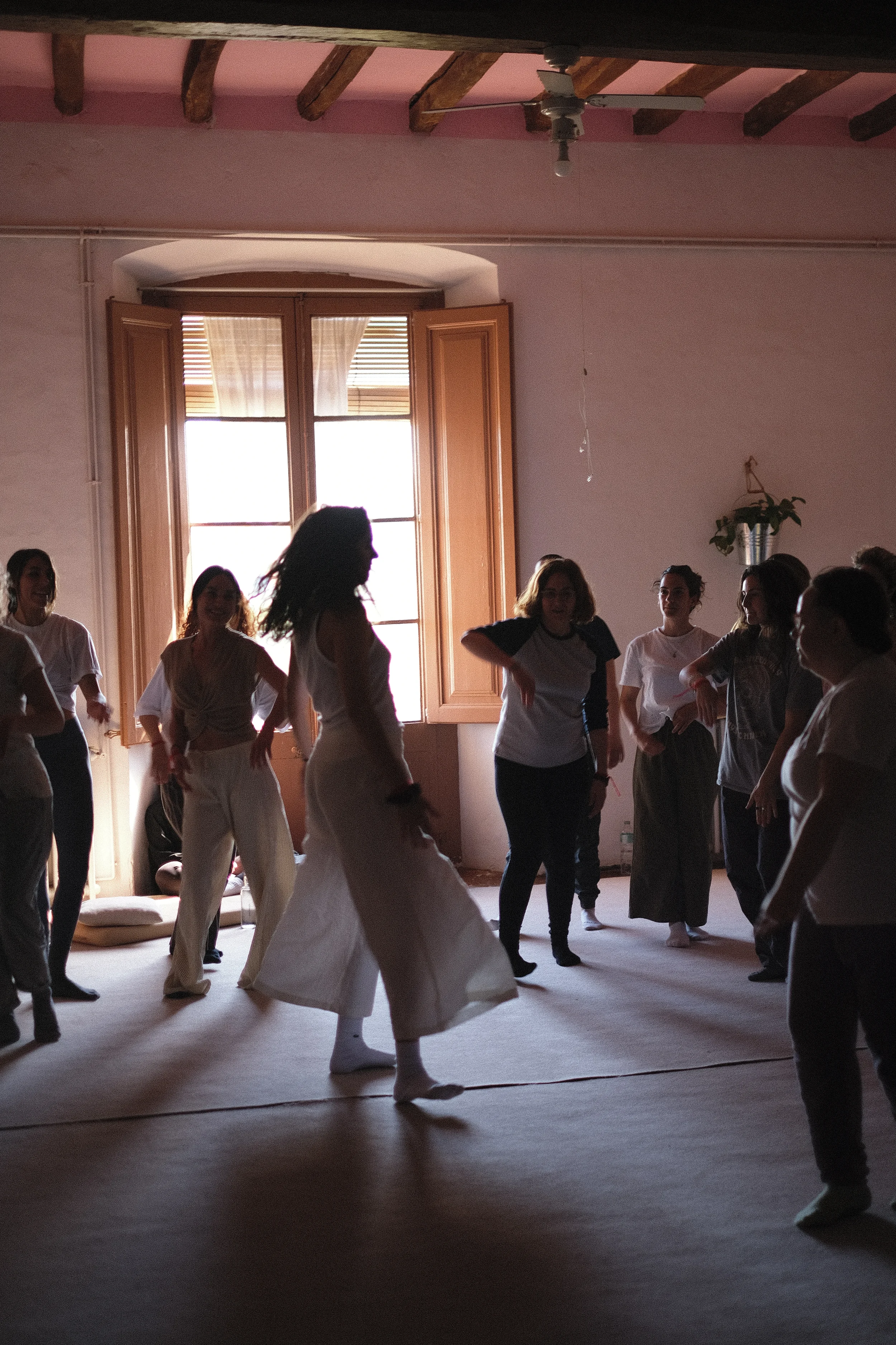 Personas bailando en un salón con ventanas abiertas y cortinas blancas, luz natural filtrándose, paredes rosadas y suelo de madera.