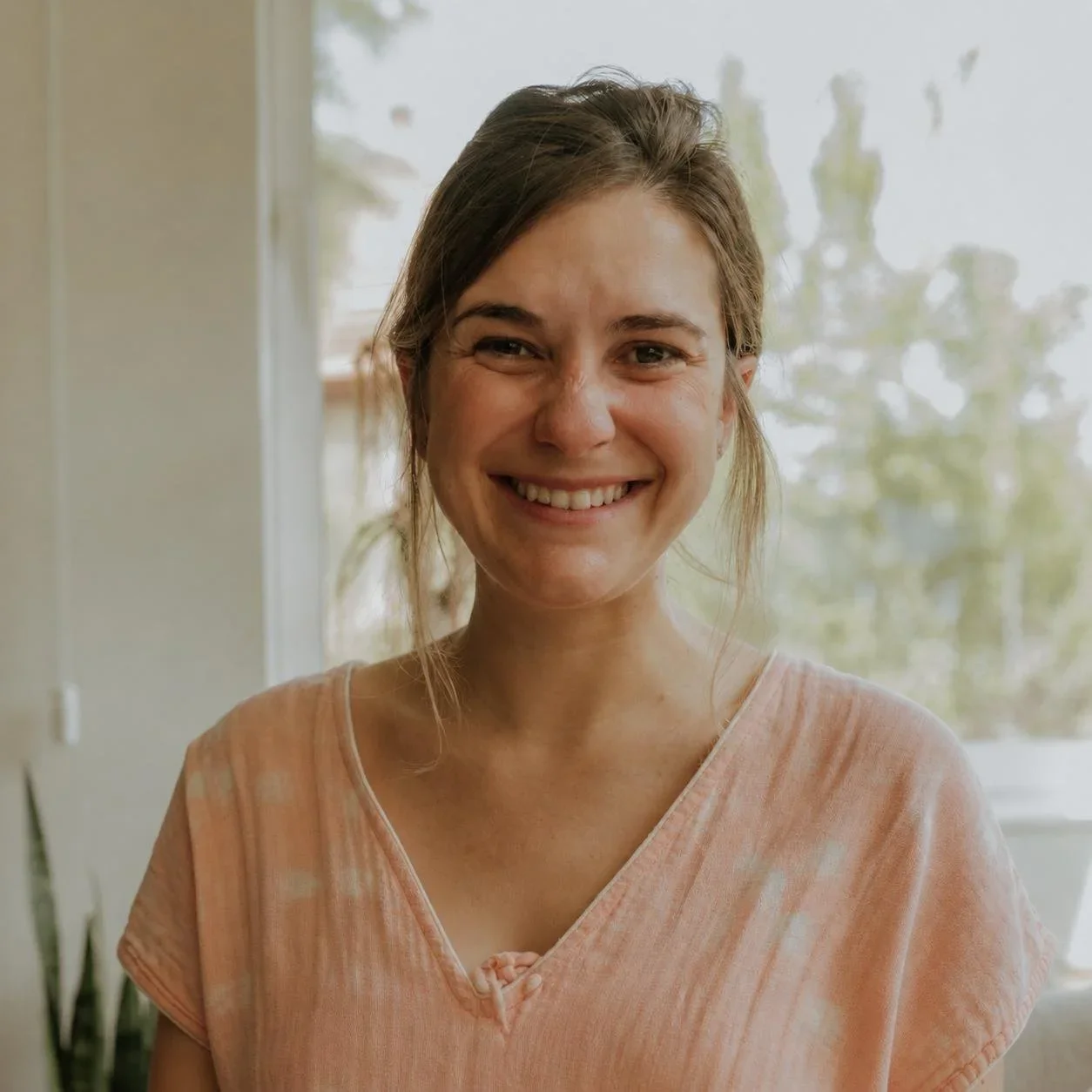 Mujer sonriendo con cabello castaño y blusa rosa claro en un espacio interior con ventanas y plantas de fondo.