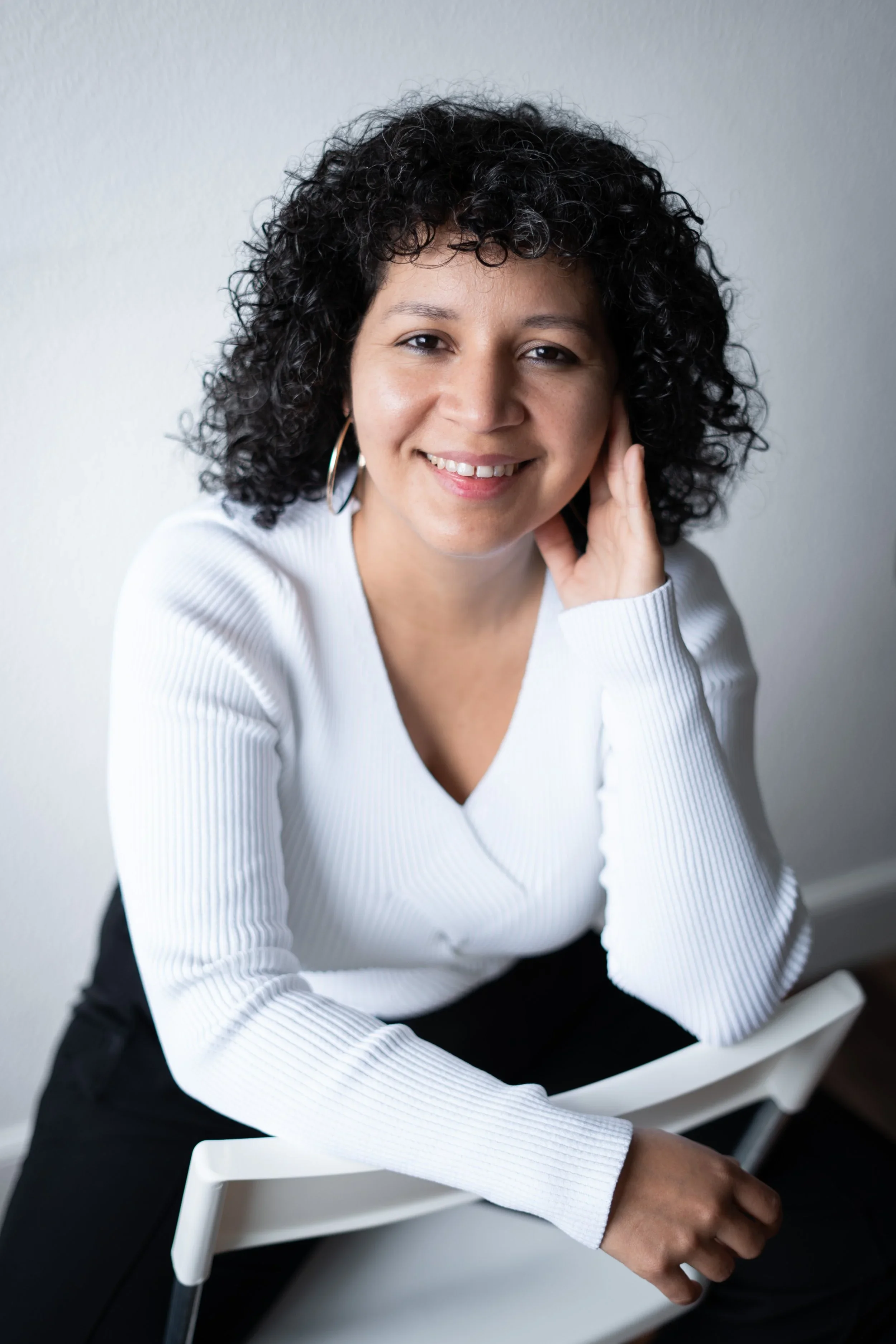 Mujer con cabello rizado negro, usando joyería de aro y camiseta blanca, sonriendo y tocándose la mejilla, sentada en una silla blanca contra una pared blanca.