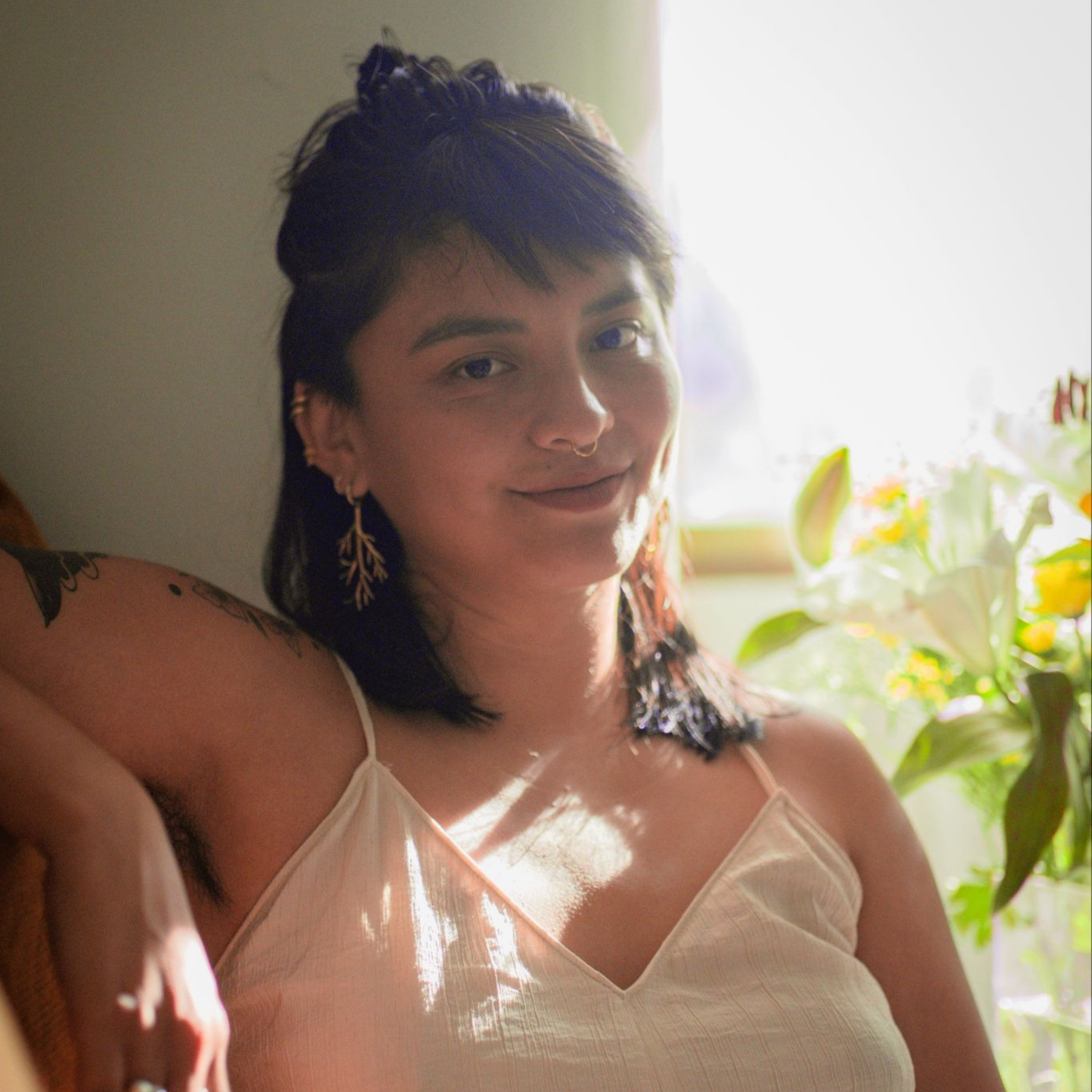 Mujer de cabello corto, con cejas delgadas, usando pendientes dorados y un vestido blanco con tirantes finos, en un entorno con luz natural y plantas verdes de fondo.