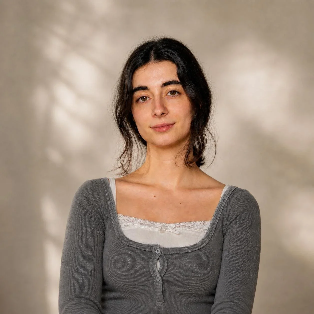 Retrato de una mujer joven con cabello oscuro y rizado, vistiendo una camiseta gris con escote entrelazado, sonriendo suavemente, fondo neutro con luz suave.
