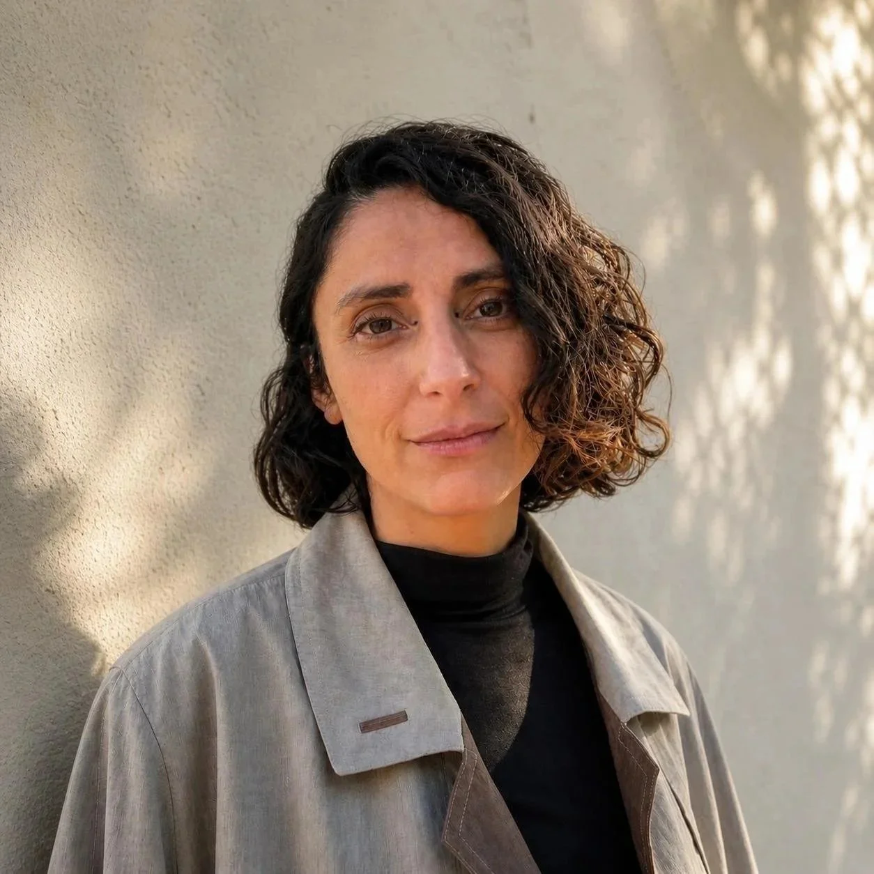 Retrato de una mujer con cabello rizado y oscuro, vestida con una chaqueta beige y camiseta negra, posando frente a una pared con sombras de luz.