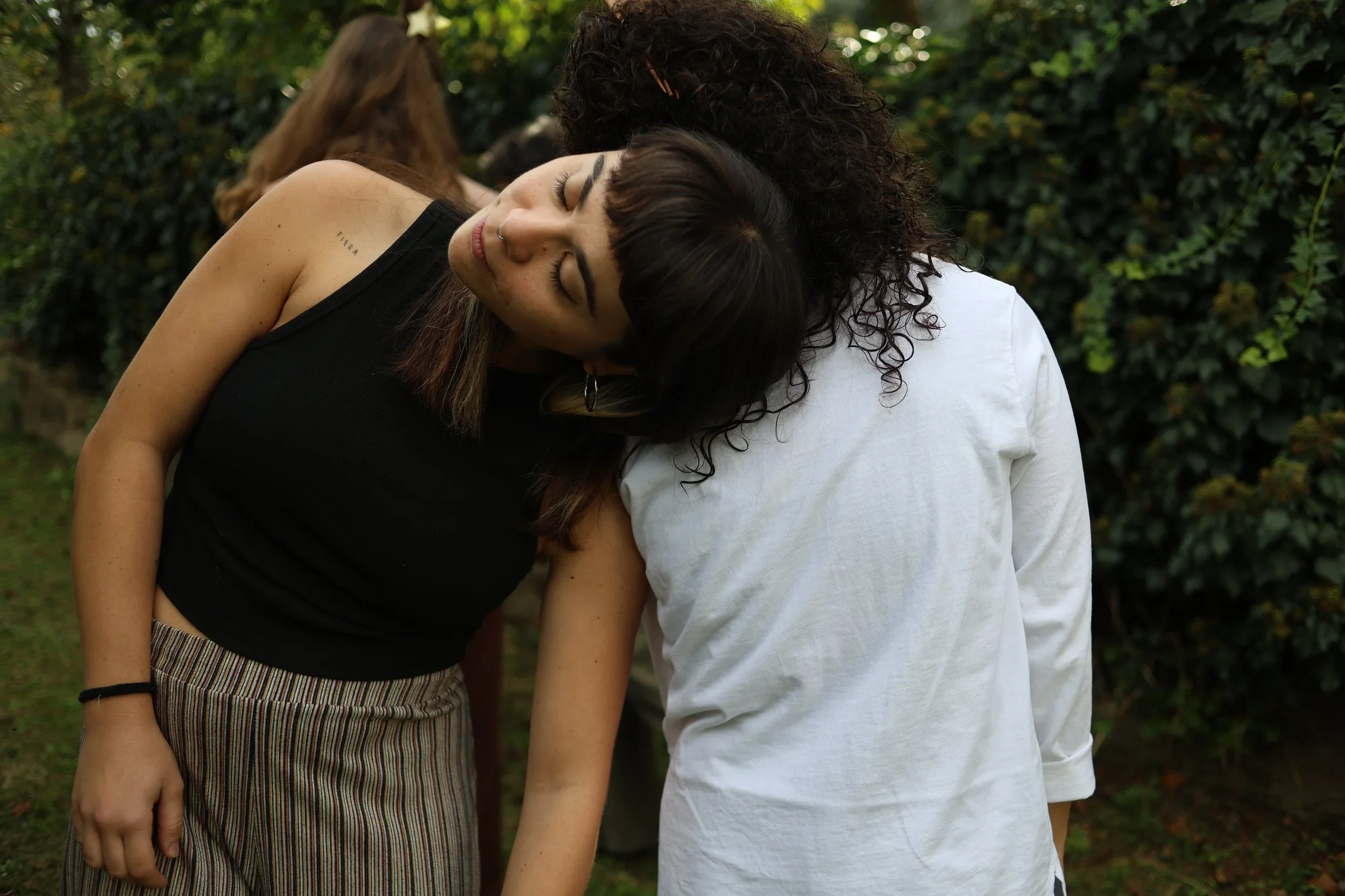 Dos mujeres con cabello oscuro abrazándose y descansando sus cabezas juntas en un entorno exterior con árboles y follaje verde.