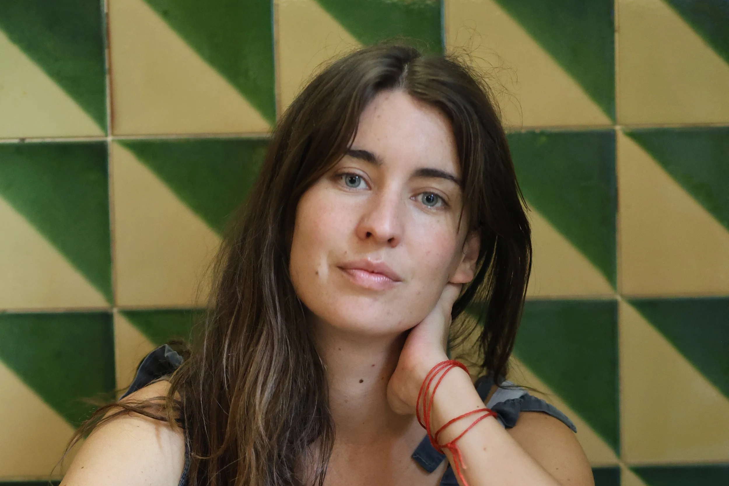 Retrato de una joven con cabello largo y arrugada, con piel clara y ojos azules, tocando su mejilla con la mano derecha, frente a un fondo de azulejos verdes y beige en patrón de triángulos.