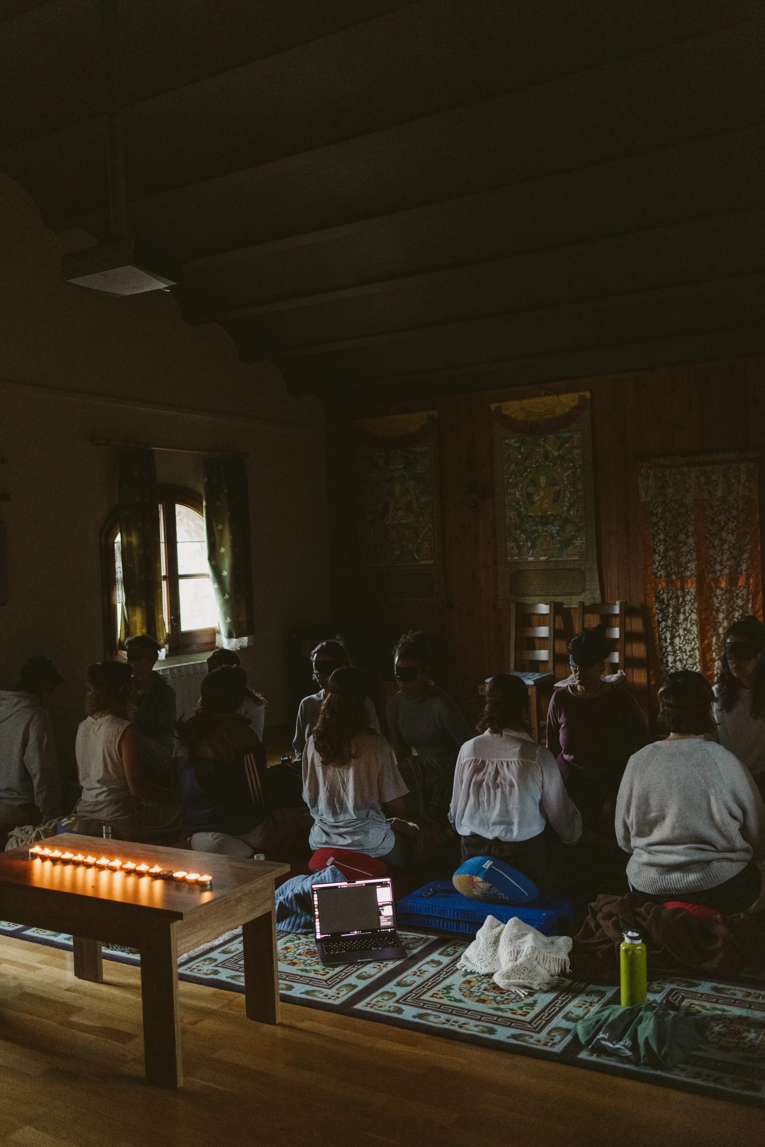 Grupo de personas participando en una actividad de meditación o oración en un espacio interior con decoración de madera y tapices en las paredes, luz suave a través de ventanas y candelas en una mesa cercana.