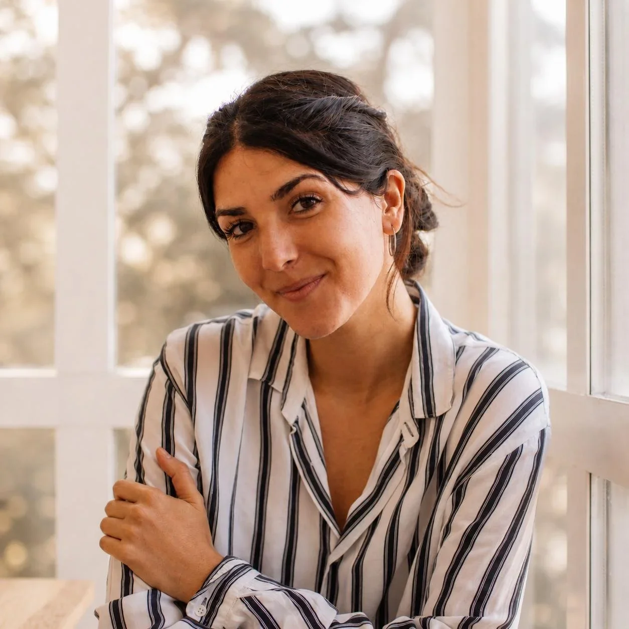 Mujer sonriendo, de pelo oscuro y peinado en bobo, viste blusa de rayas blancas y negras, junto a ventana con vista a árboles.