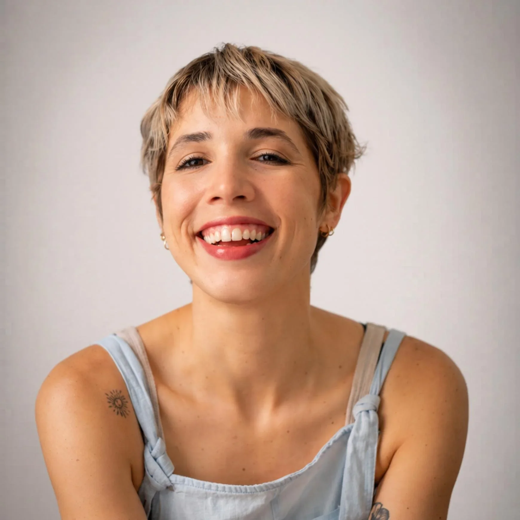 Mujer con cabello corto, rubio y con tatuajes en los brazos, sonriendo y usando un vestido sin mangas de color gris claro con detalles en azul.