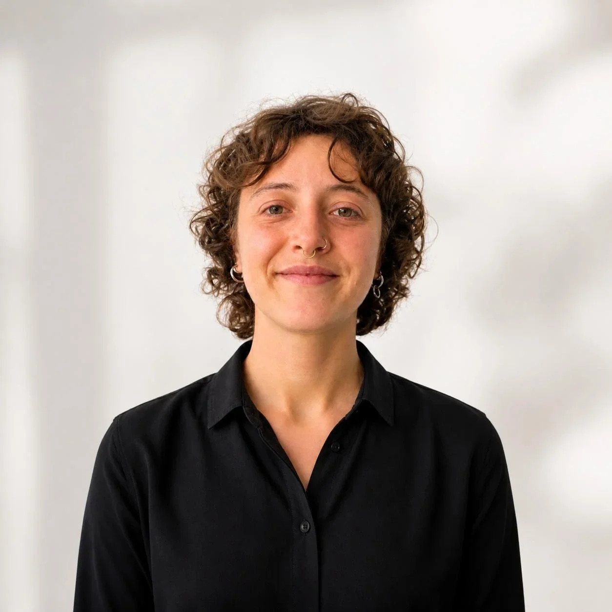 Retrato de una mujer con cabello rizado y oscuro, sonriendo ligeramente, lleva una blusa negra y está de frente en un fondo blanco.