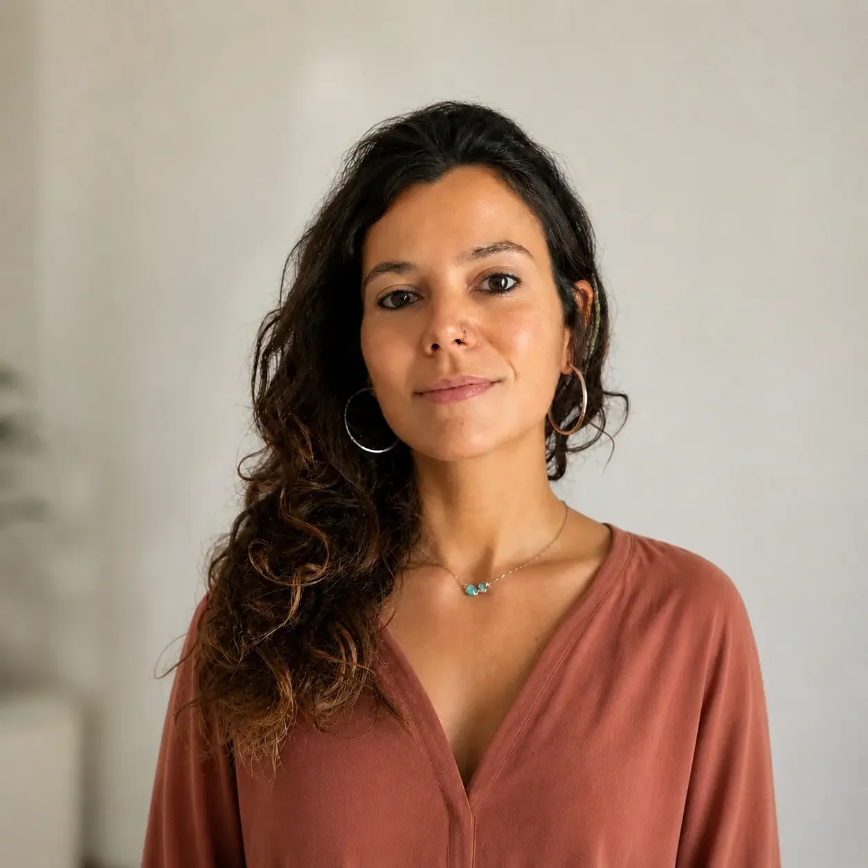 Retrato de una mujer con cabello rizado, usando un anillo, aretes grandes y un collar con piedras azules, con una pared clara de fondo.