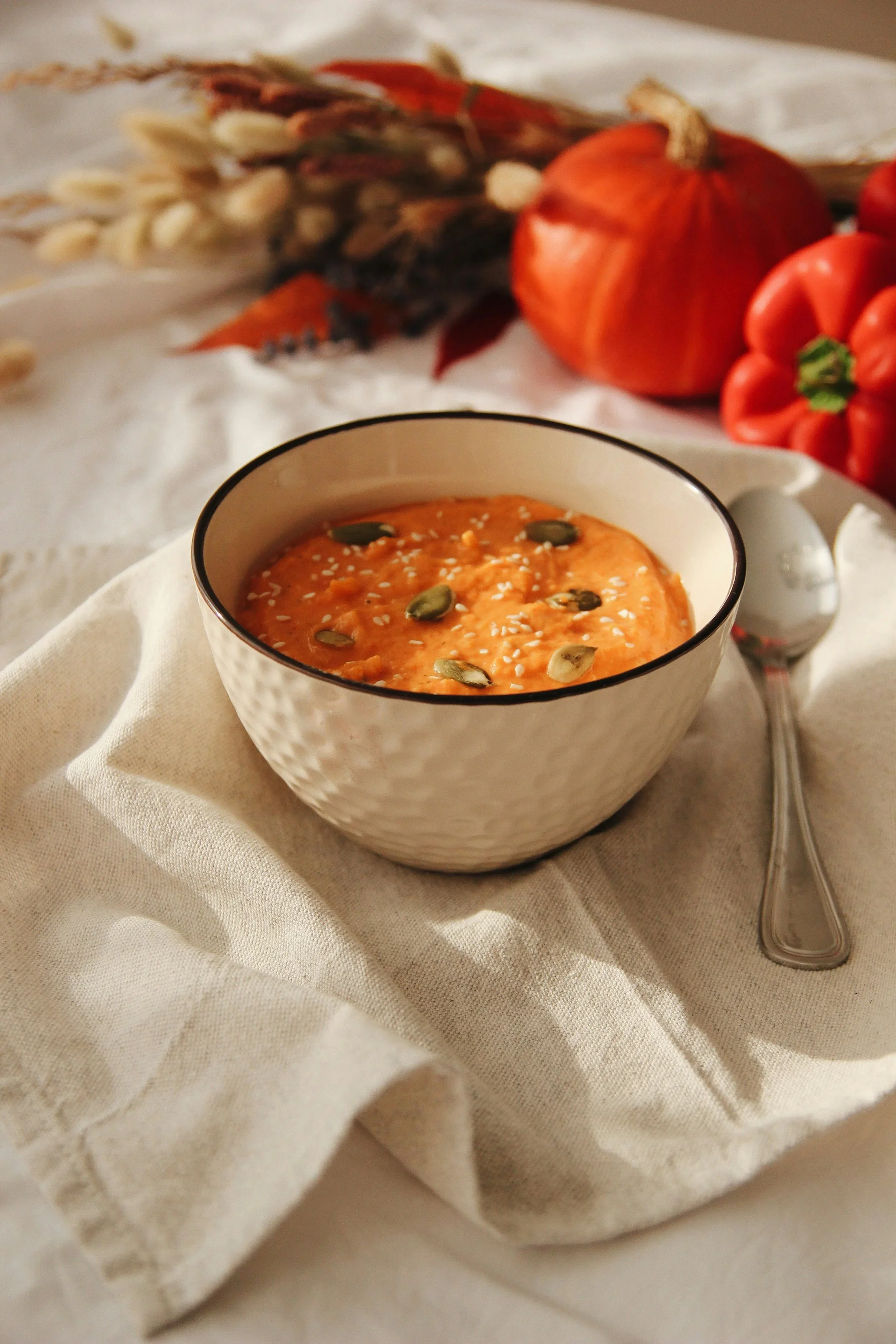 Una taza blanca con borde negro con sopa de tomate decorada con semillas de calabaza y sésamo, sobre una tela blanca, con una cuchara de metal a su lado y fondo difuso de vegetales y flores.