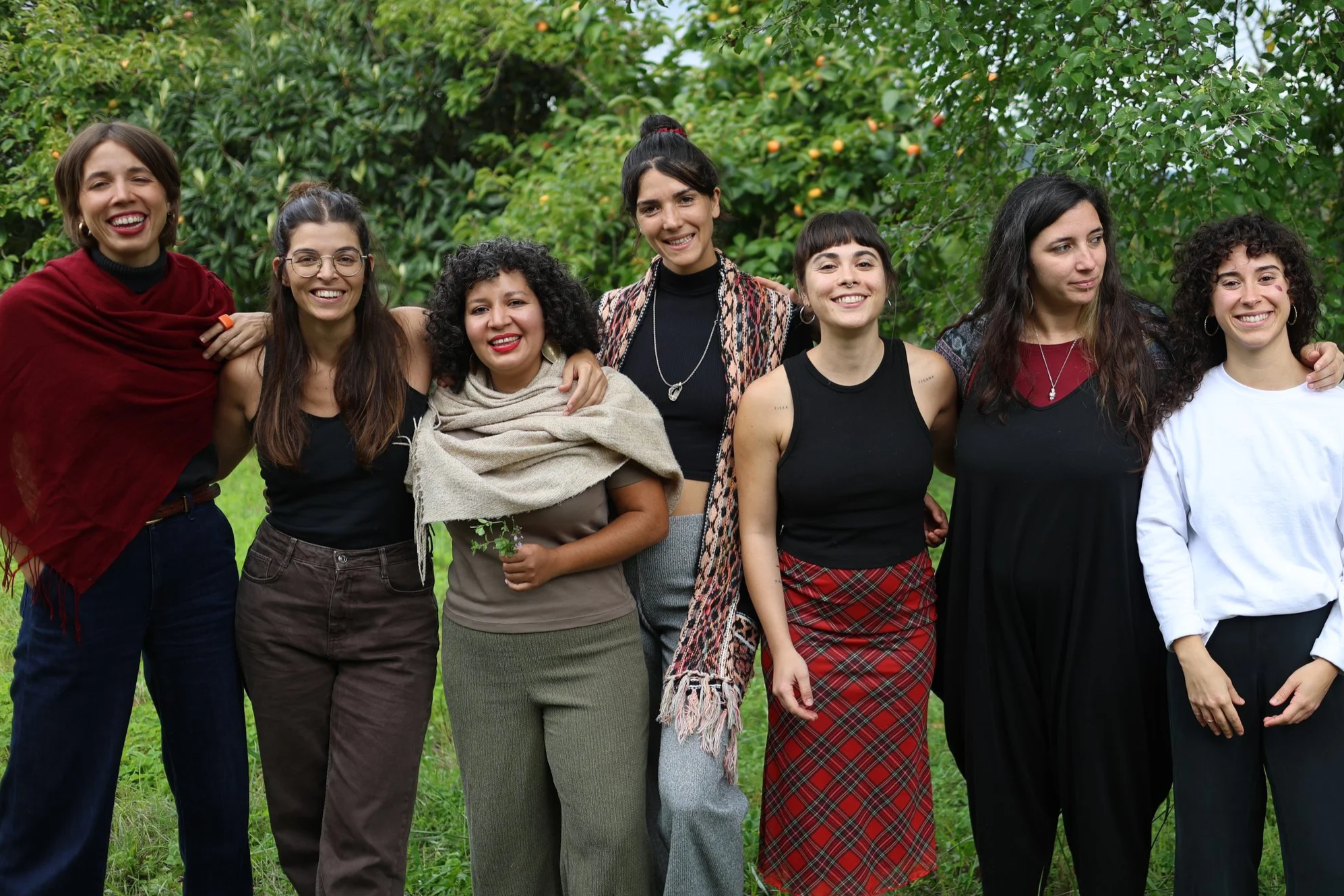 Grupo de mujeres jóvenes, algunas con brazos alrededor, sonriendo y posando al aire libre con árboles y naranjos de fondo.