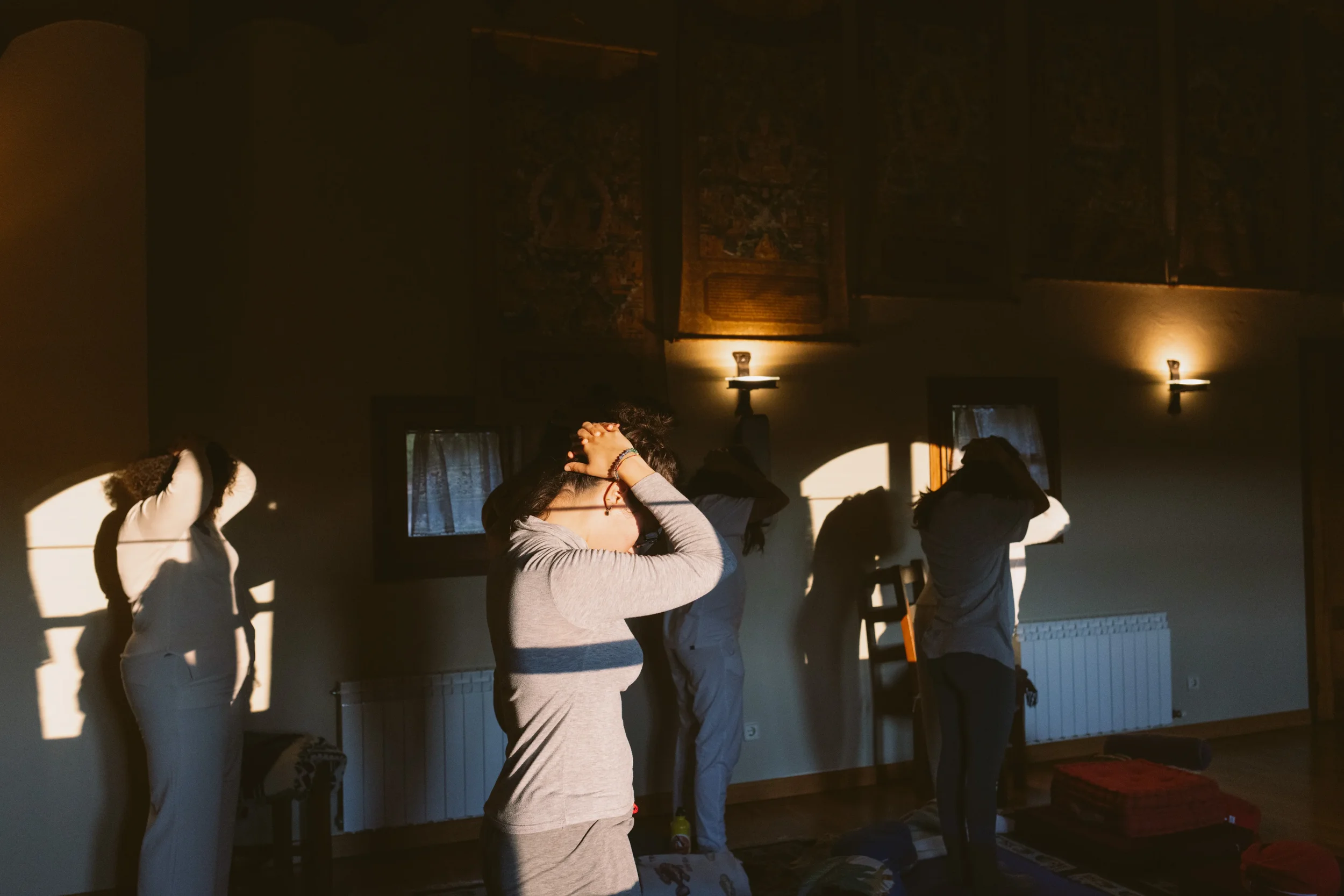 Varias personas en una sala recibiendo una sesión de meditación o yoga, con luz de sol iluminando la habitación y cuadros en la pared.