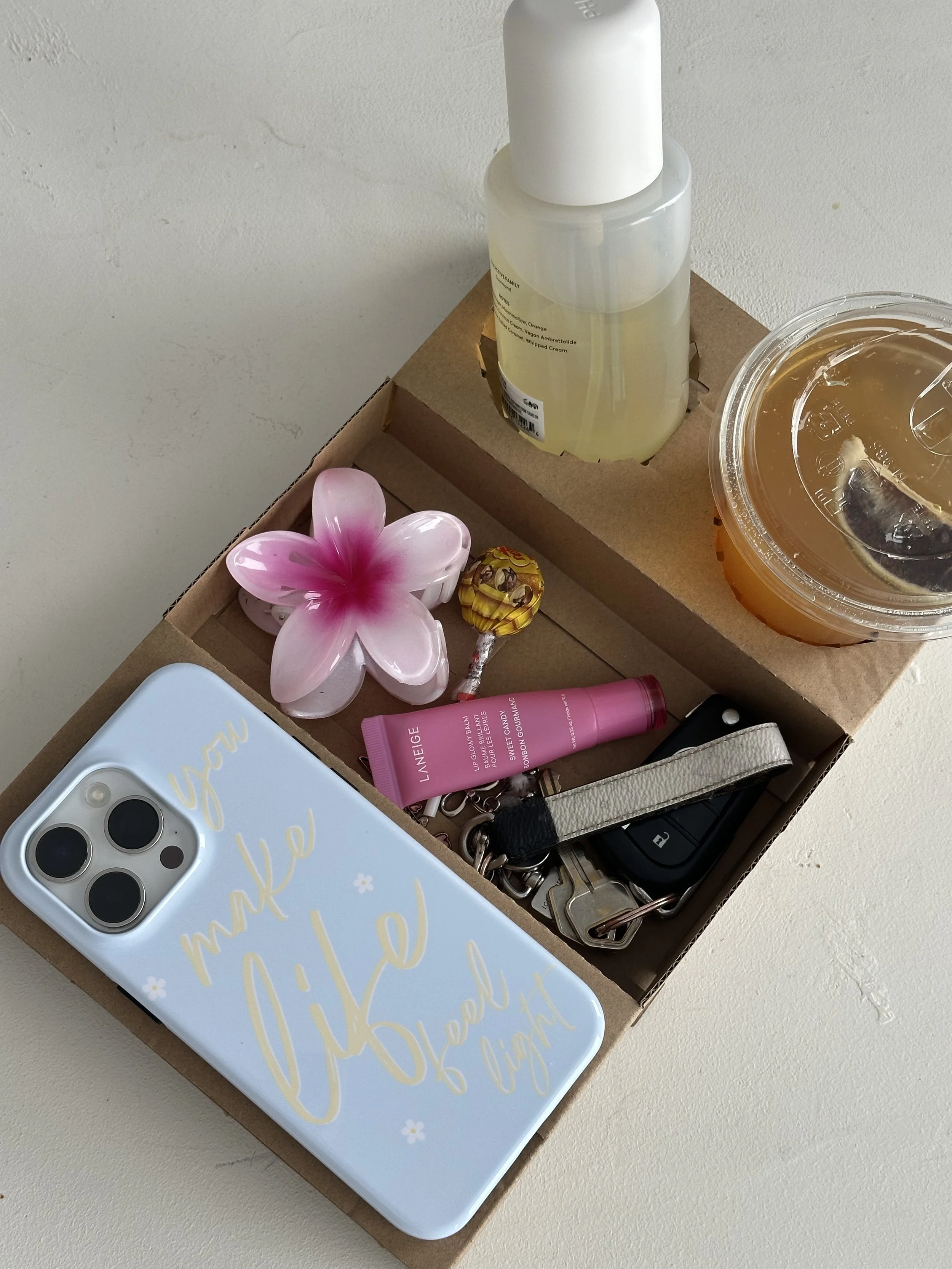 A cardboard box containing a pink flower-shaped soap, a tube of pink lip balm, a set of keys with a black and white strap, a keychain with a yellow bee, a white and beige phone case , a bottle of lotion, a container of iced tea, and a small bottle of yellow liquid.